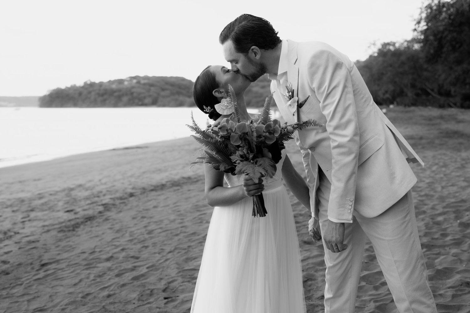 Romantic beach wedding couple embracing during sunset with tropical flowers and ocean backdrop.