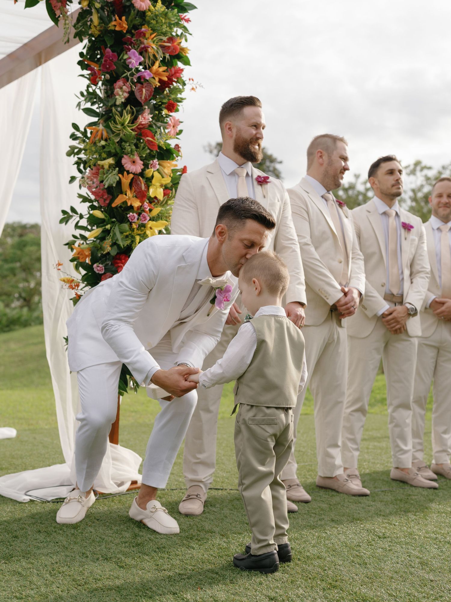 Groomsmen in white suits posing playfully on grass near floral wedding arch.