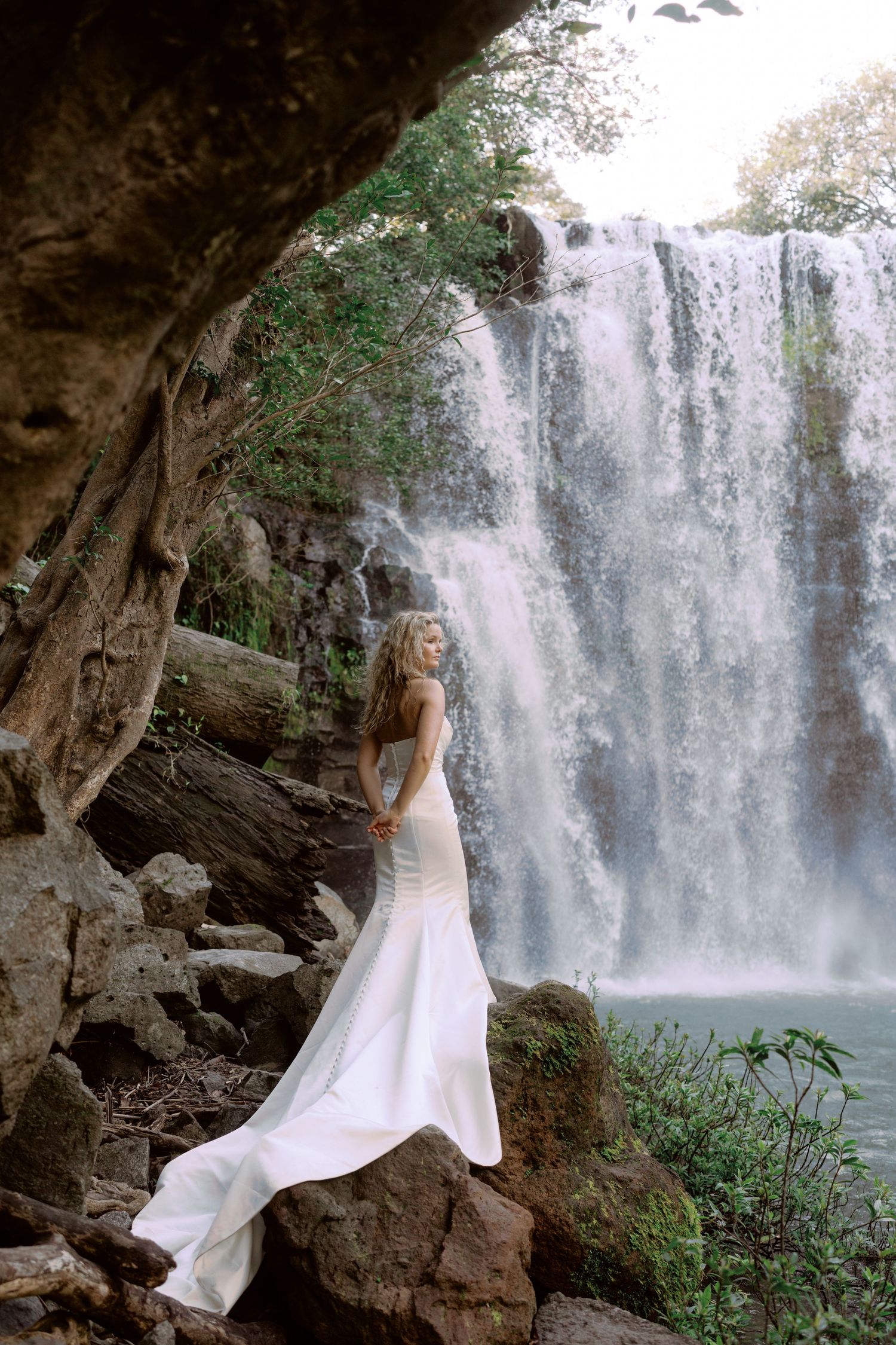 A white dress flows gracefully against the backdrop of a majestic waterfall in a scenic natural setting.