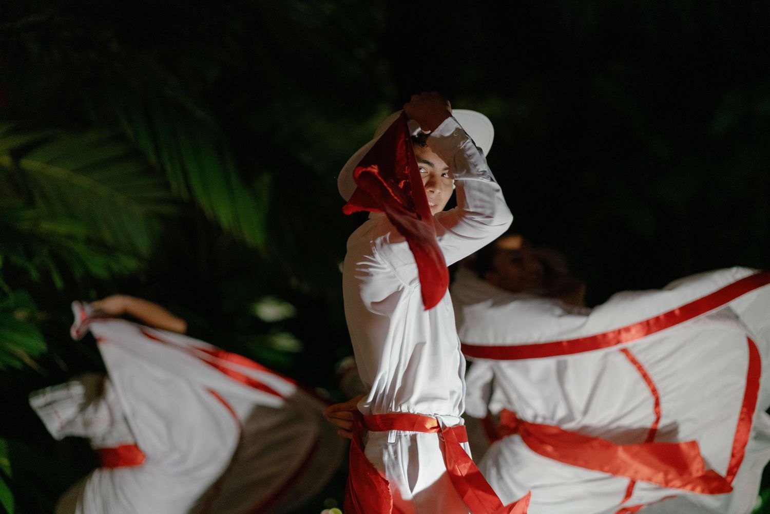 Dancers in white costumes with red accents perform dynamic movements in low light.