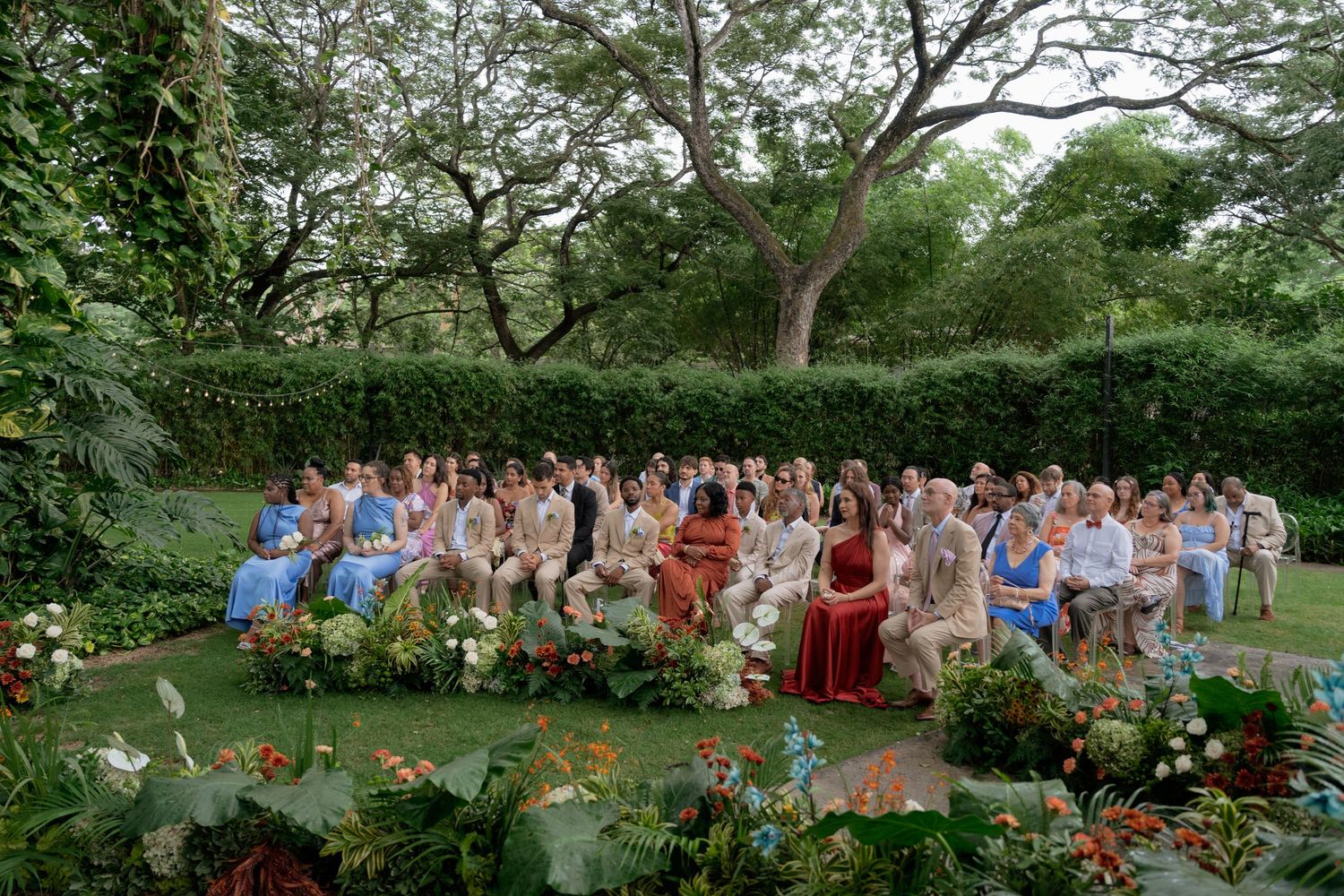 Large group photo of wedding guests gathered outdoors in a garden setting.