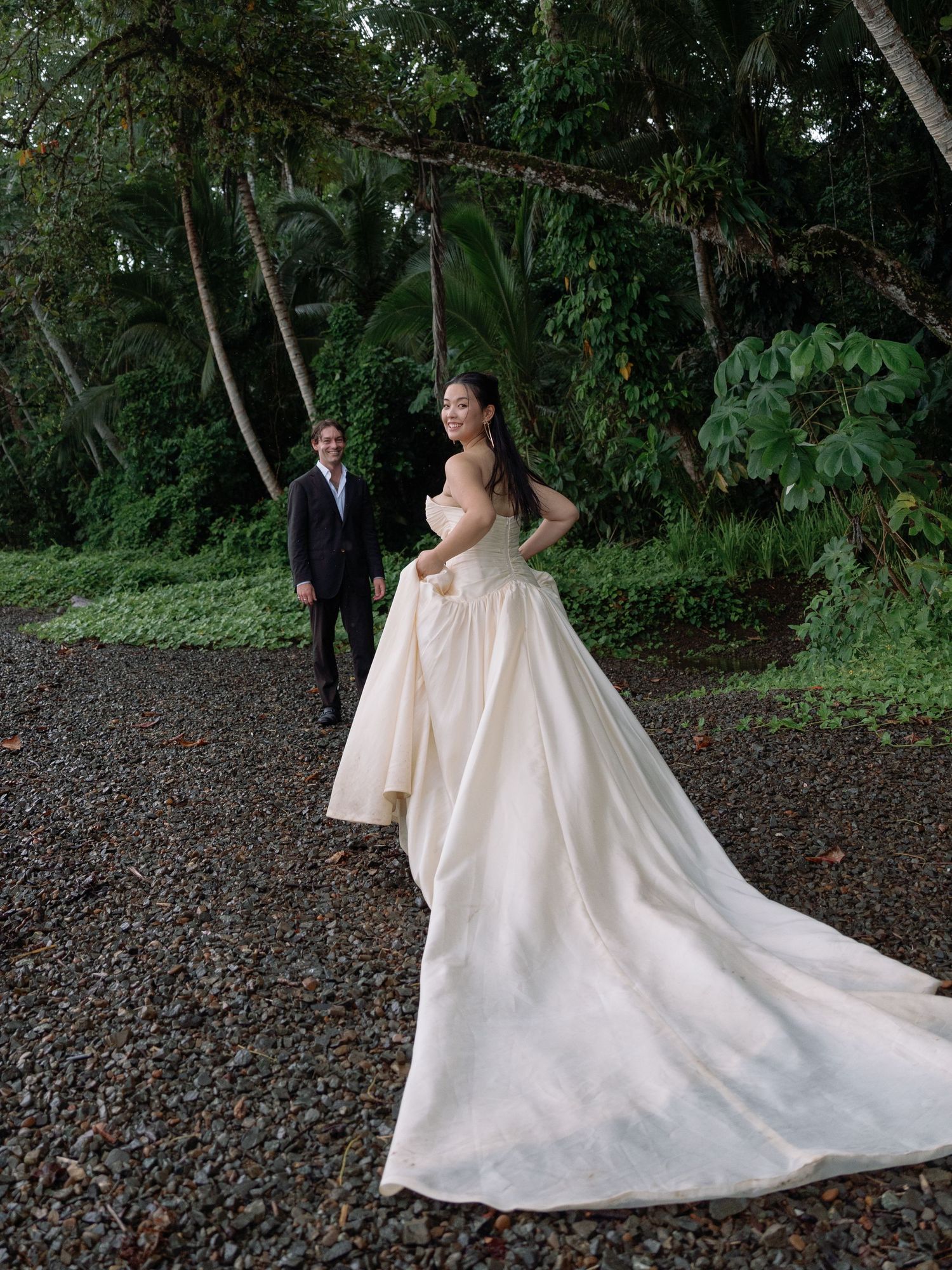 A bride in a flowing train gown poses on a gravel path with tropical greenery.