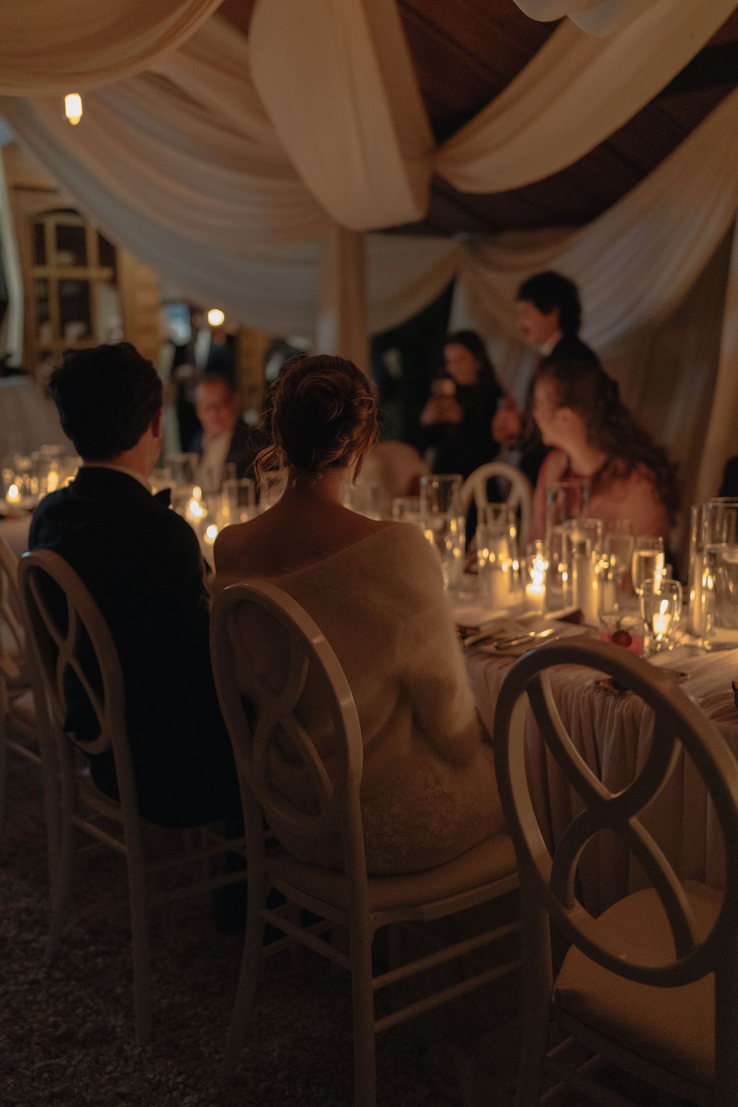 Bride and groom seated at candlelit wedding reception table in elegant draped tent venue.