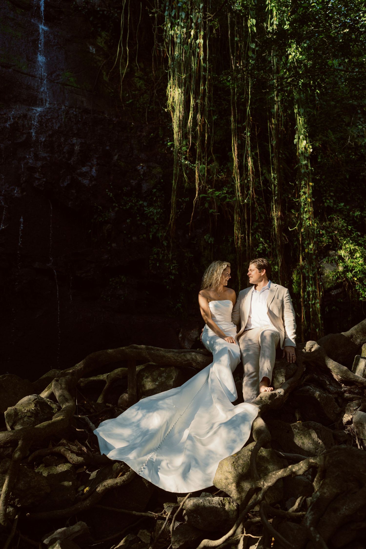 A couple in formal attire poses romantically on dark rocks surrounded by lush tropical foliage.