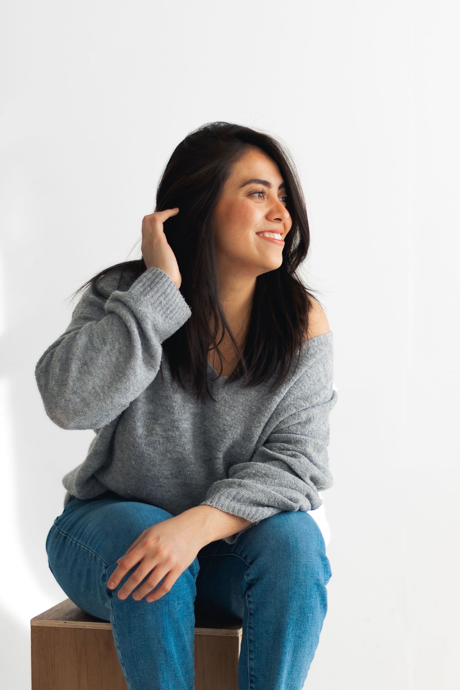 Model in cozy gray sweater and jeans sits casually on wooden block against white background.