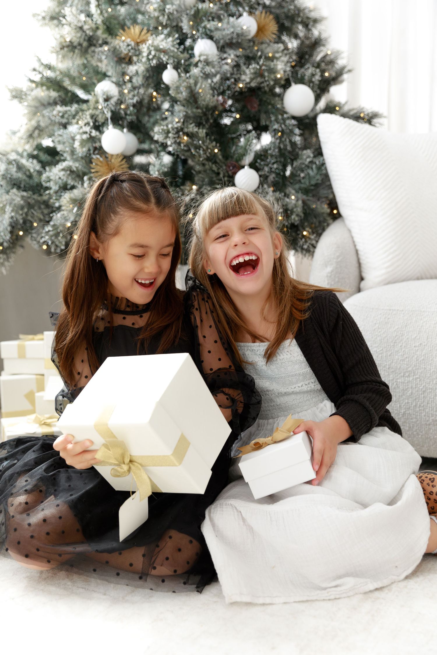 Two excited girls laughing and opening white Christmas gifts in front of a decorated silver and white Christmas tree.