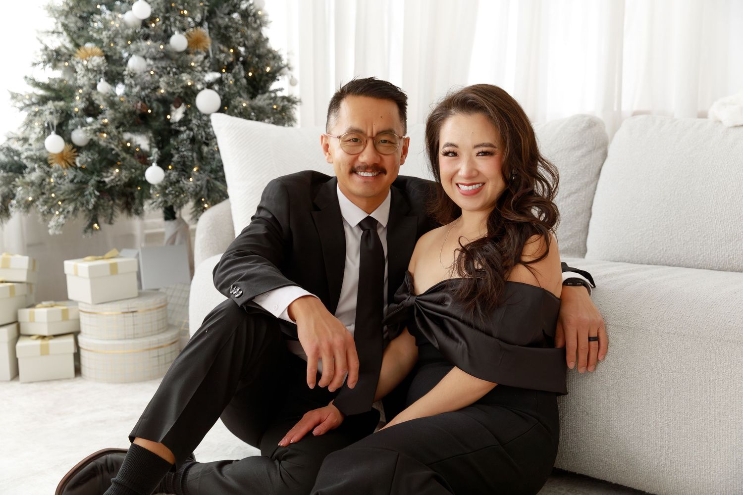 Couple poses lovingly near a decorated Christmas tree with wrapped presents in a bright white room.