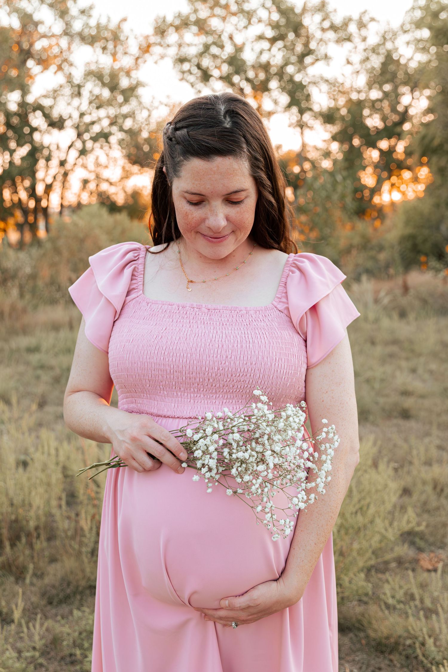 A maternity photo of someone in a pink dress holding baby's breath flowers at sunset.