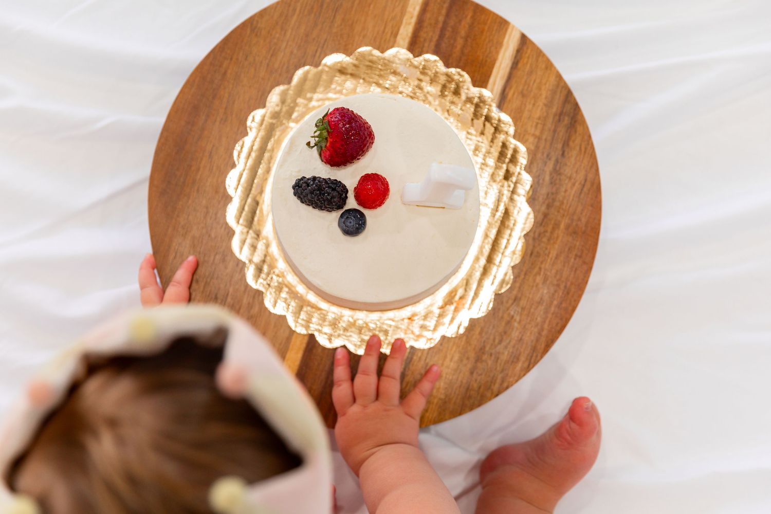 Baby reaching for a elegant white cake topped with fresh berries on a wooden board.