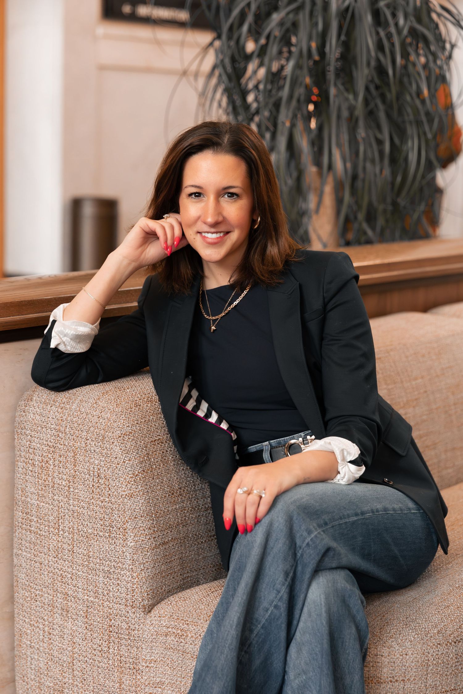 Professional woman in black blazer sits casually on beige couch in modern office setting.