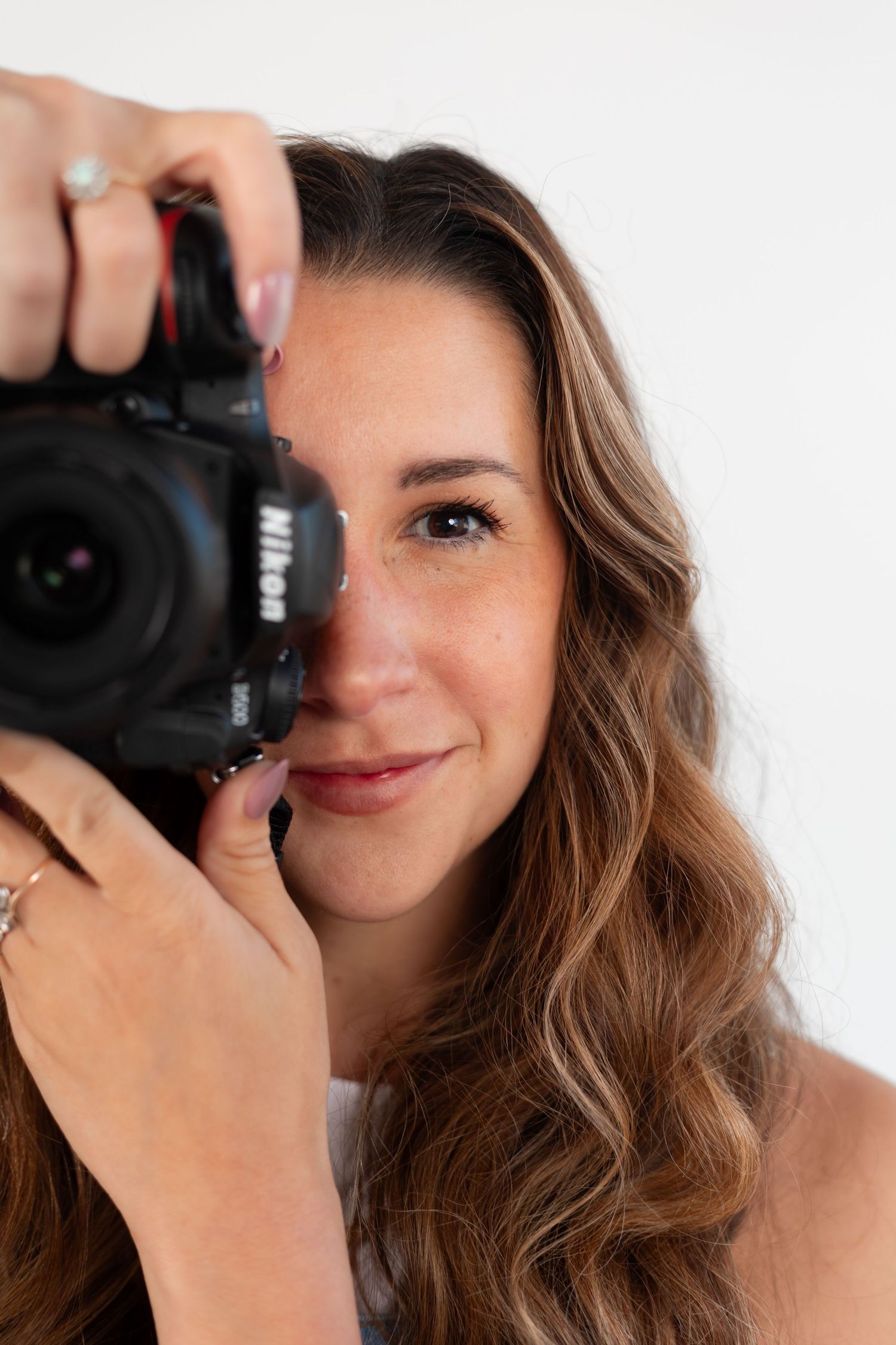 A photographer with wavy hair holds a professional camera against a white background.