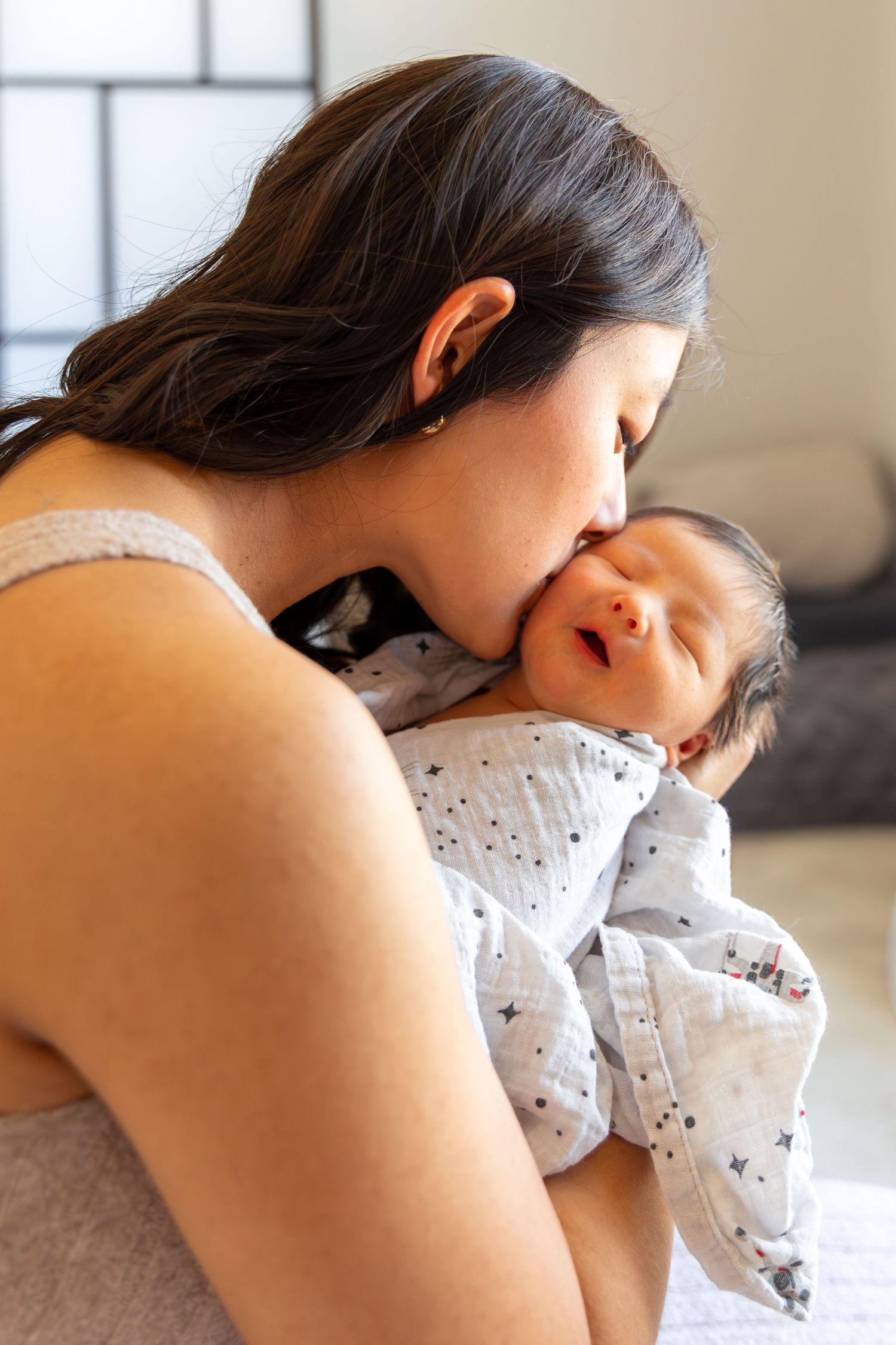 A mother tenderly kisses her sleeping newborn baby wrapped in a white muslin swaddle blanket.