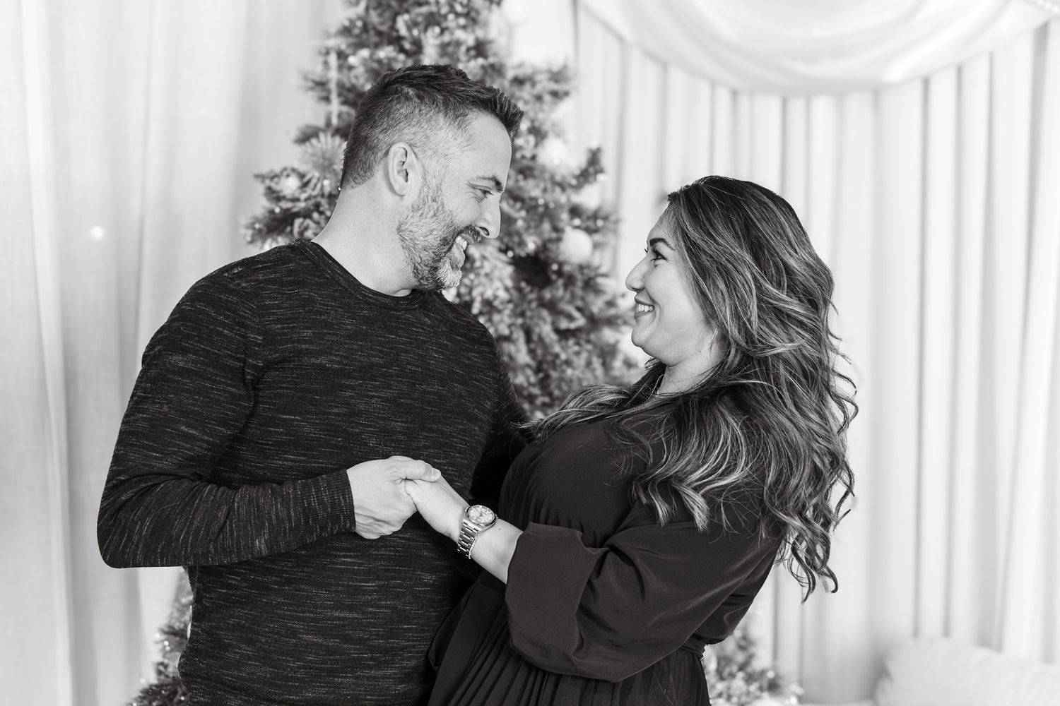 Black and white photo of a smiling couple holding hands in front of a Christmas tree.