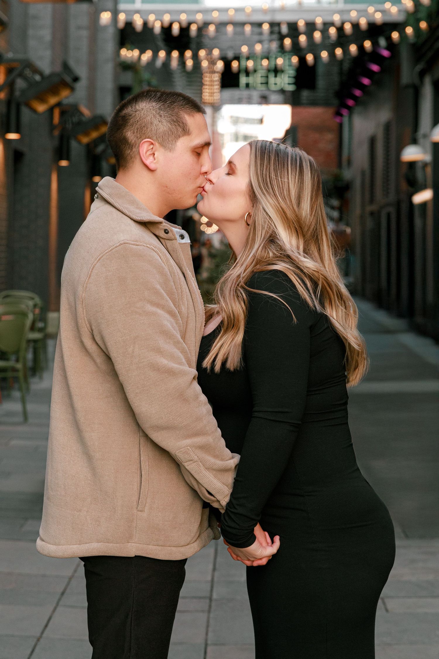 Romantic couple sharing a kiss under string lights in an urban alley setting.
