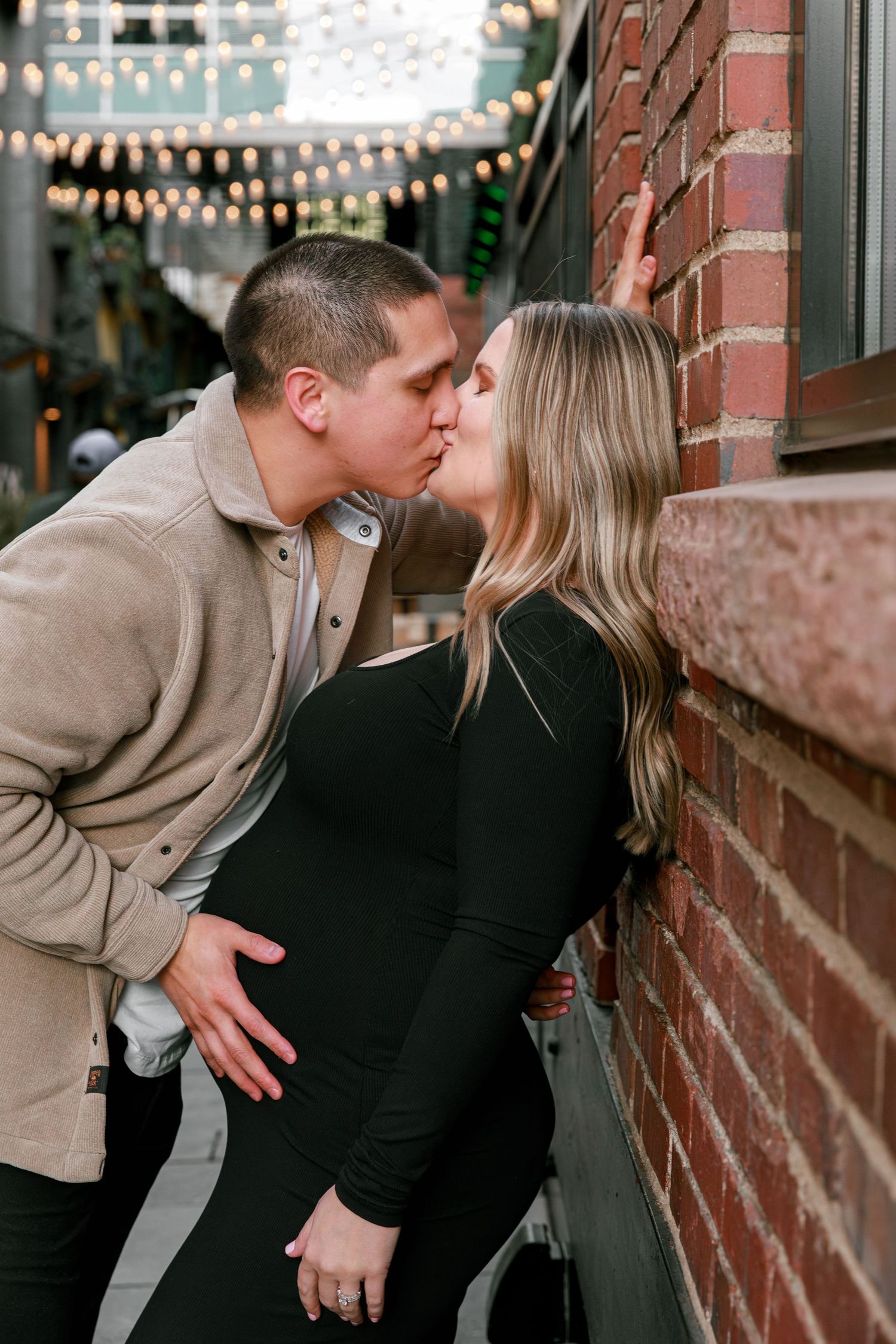 Romantic couple sharing intimate moments against a brick wall with string lights.