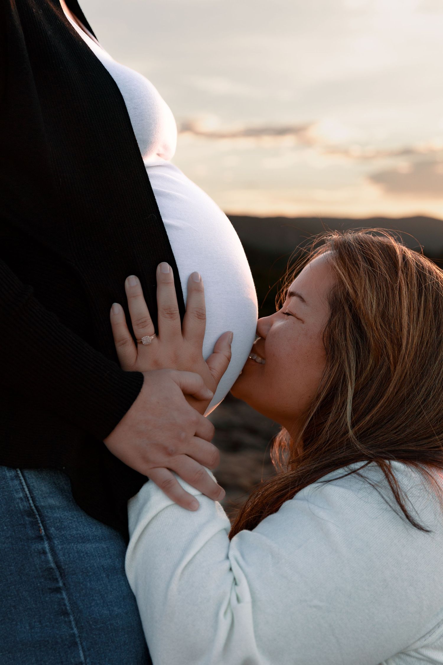 Intimate maternity photo of expectant mother kissing pregnant belly during sunset.