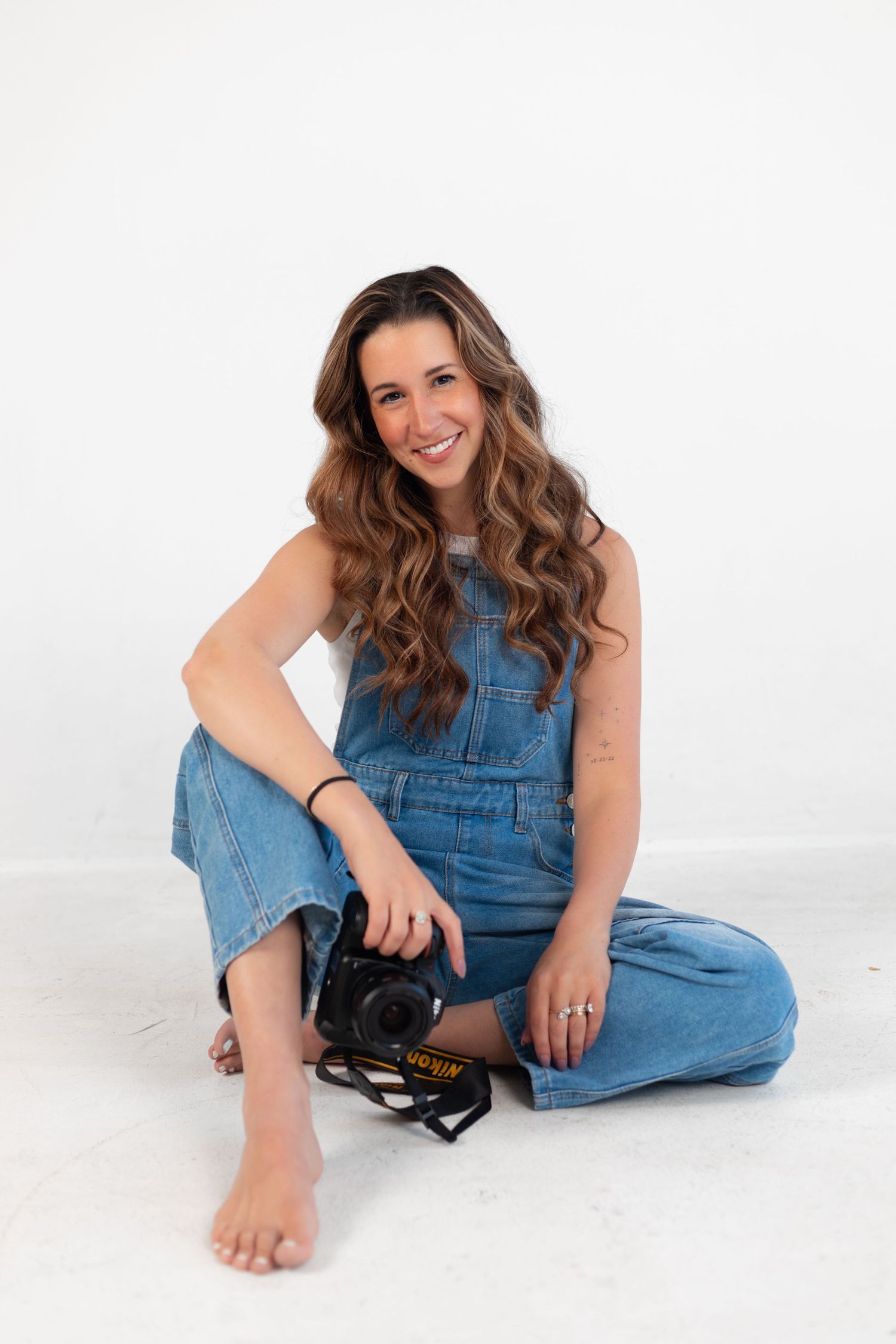 Woman in denim overalls sits casually with camera on white background.