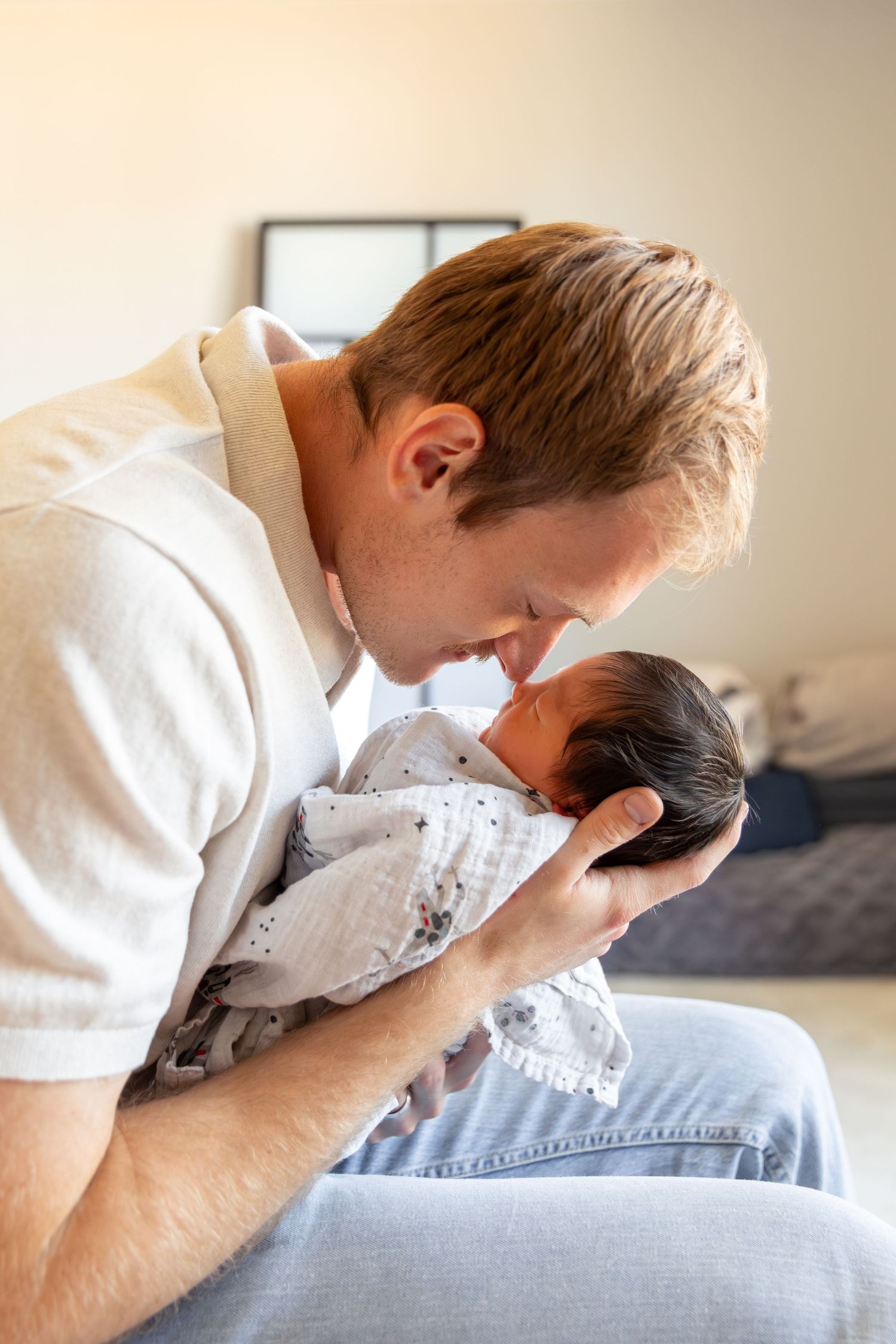 A father tenderly touches noses with his swaddled newborn baby while seated indoors.