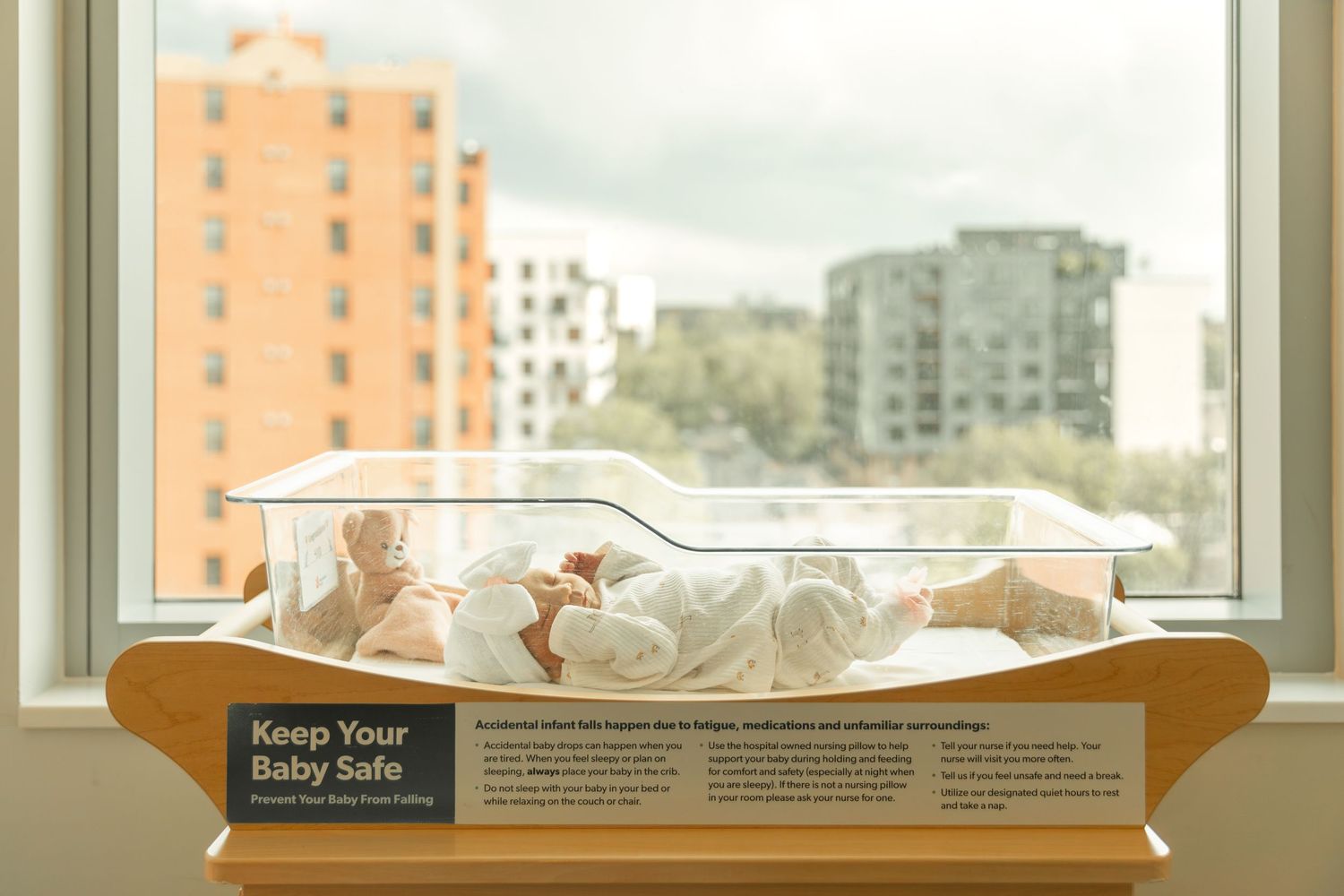 Baby sleeping safely in a clear bassinet with a Keep Your Baby Safe sign near a window.
