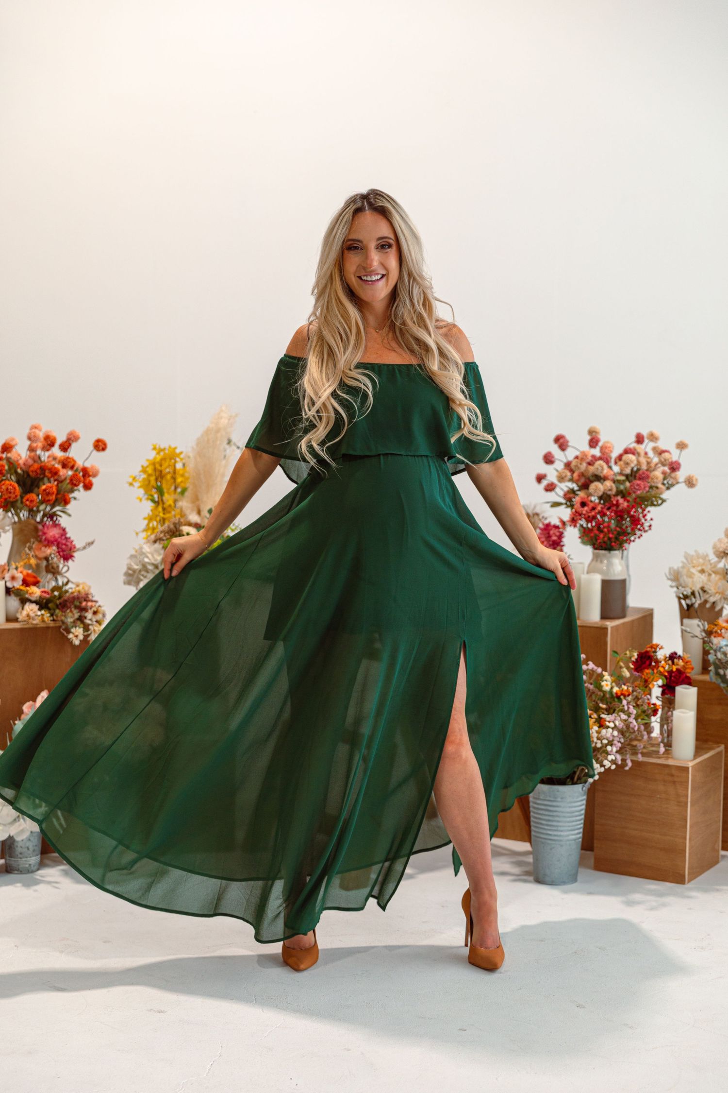 Emerald green flowing maxi dress with floral backdrop and wooden accents in studio photoshoot.
