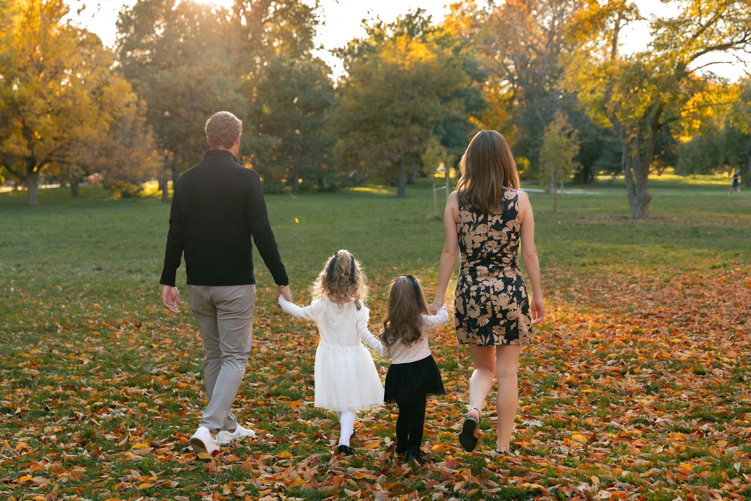 Family of four walking hand-in-hand through a sunlit autumn park with golden fallen leaves.
