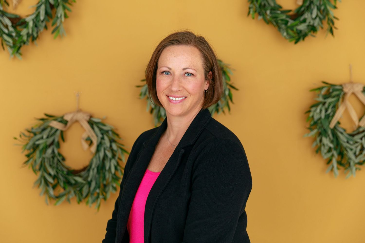 Woman in black blazer and pink top poses against yellow wall with greenery decor.