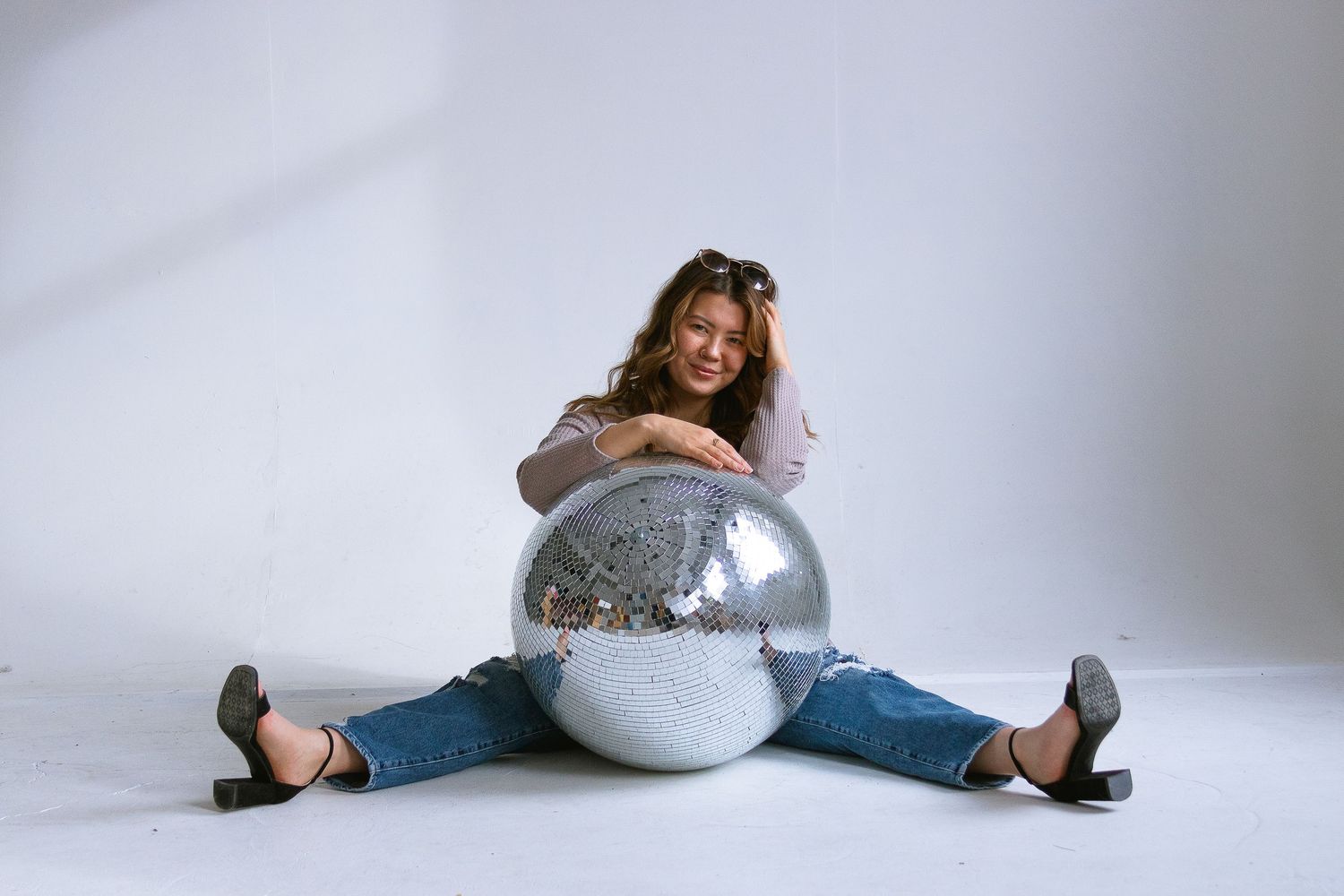 Model in casual outfit sits with legs spread around silver disco ball against white backdrop.