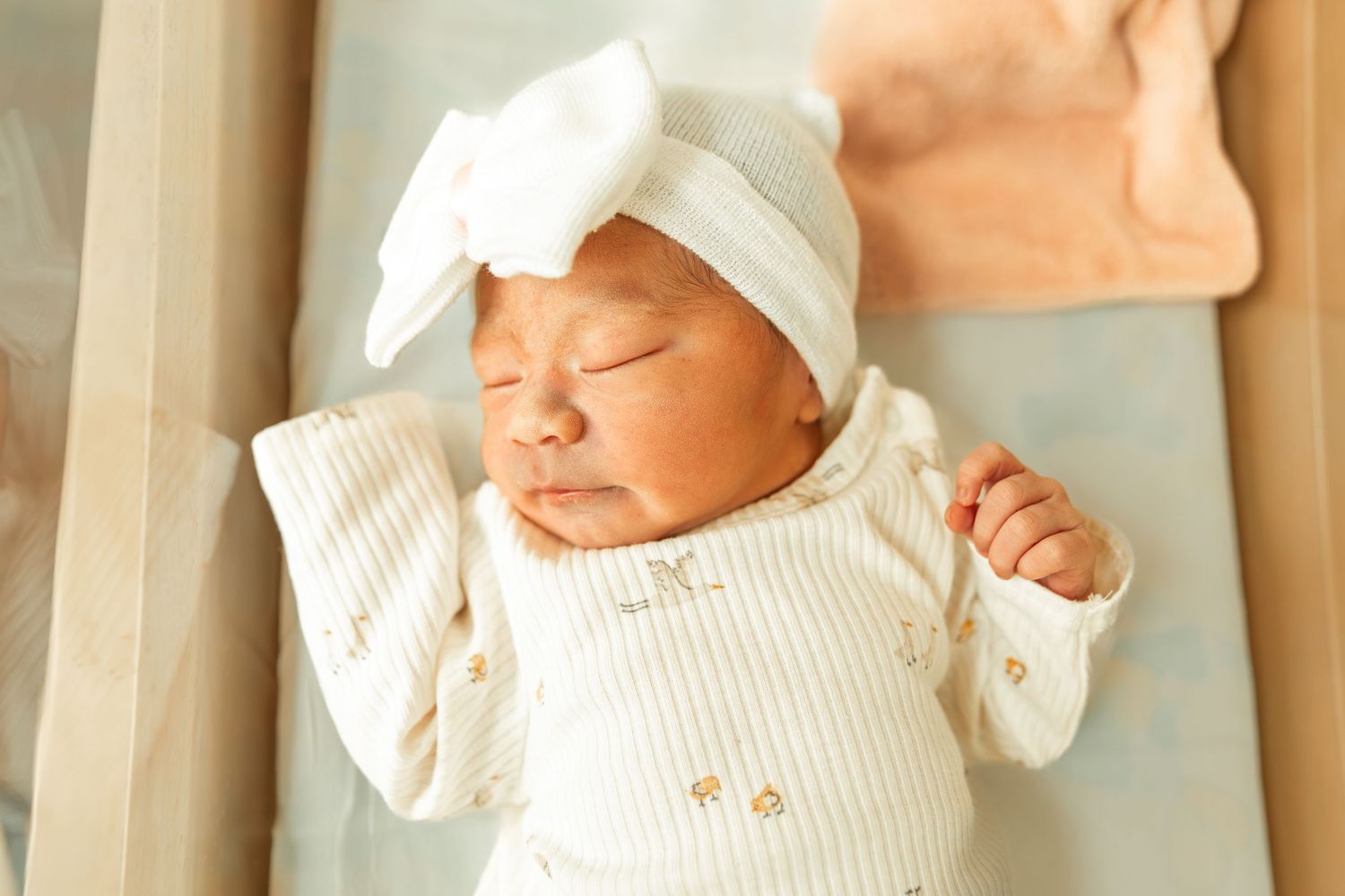 Newborn baby sleeping peacefully in a crib wearing a white onesie and bow hat.