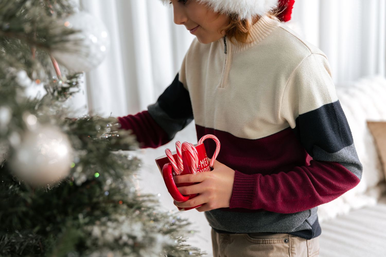Child in Santa hat holding red mug with candy canes near decorated Christmas tree.