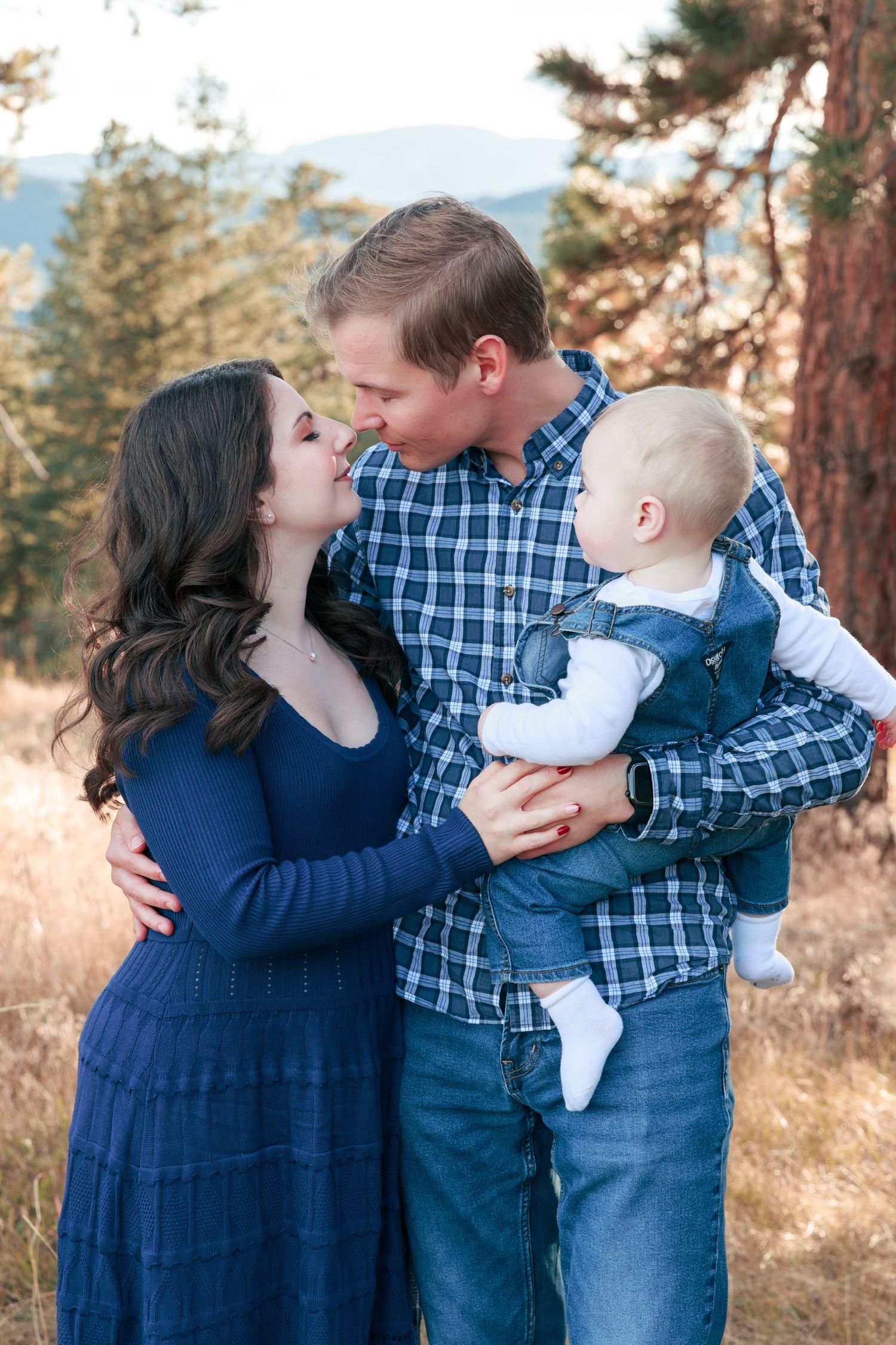 A young family shares a tender moment outdoors, with parents kissing while holding their baby in a mountain forest setting.
