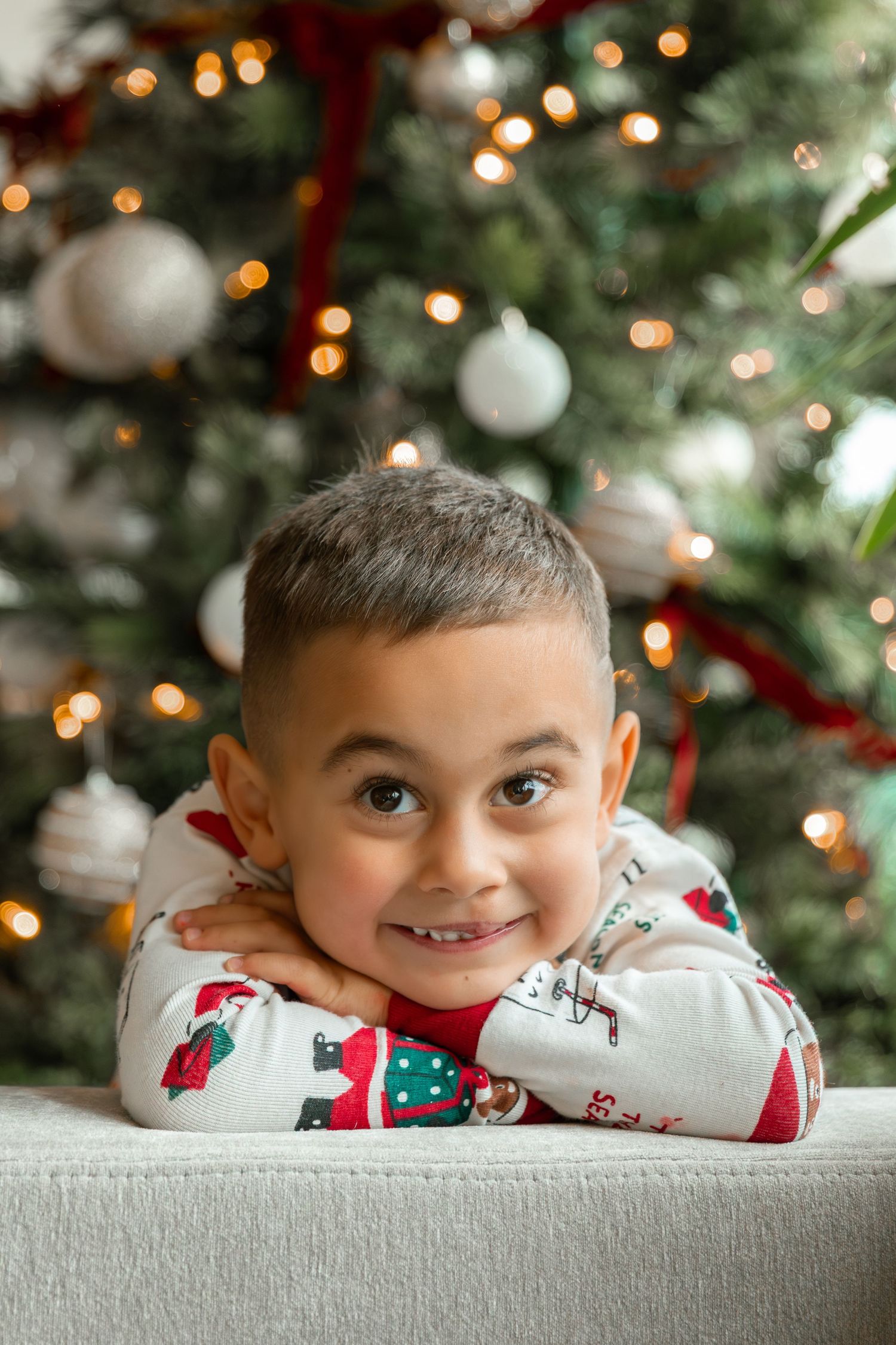 Smiling boy in Christmas pajamas resting chin on arms in front of a decorated holiday tree with lights.