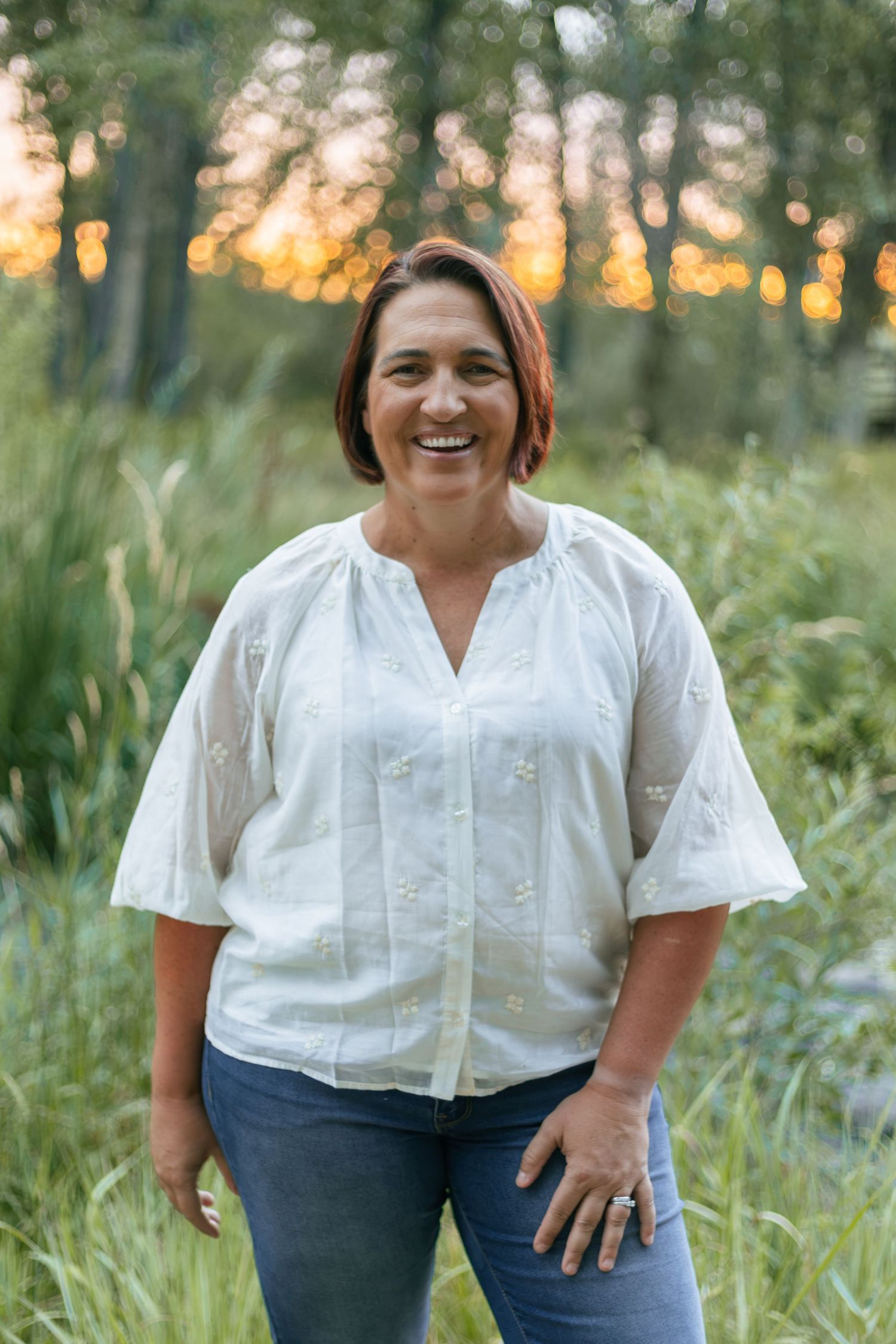 Smiling woman in white embroidered blouse and jeans standing outdoors in lush green field at sunset.