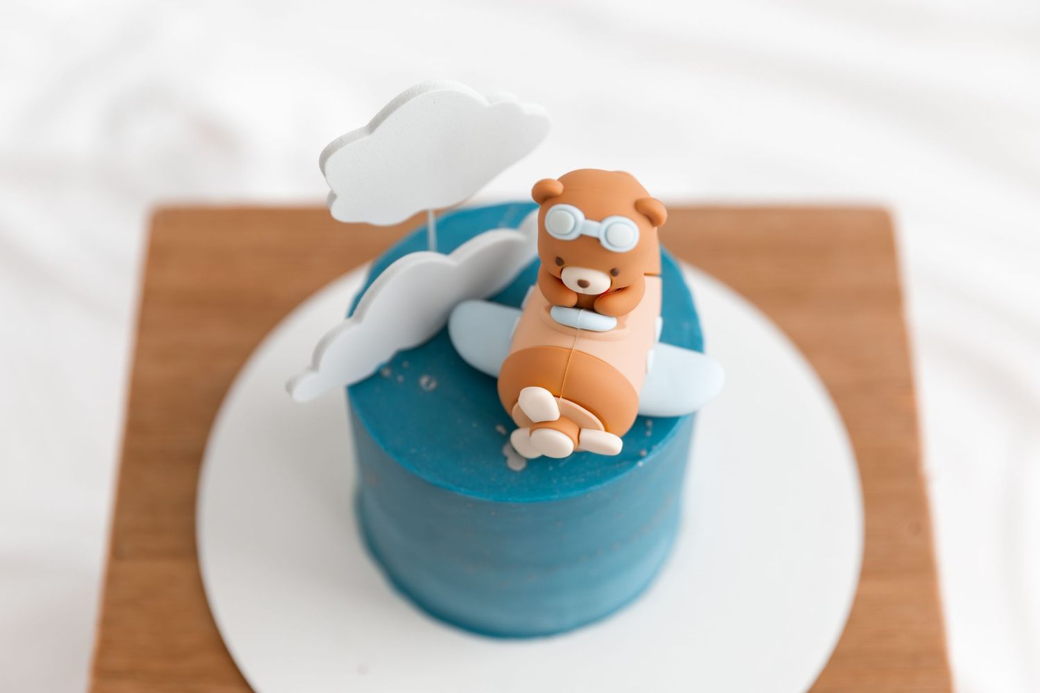 Blue fondant cake topped with a cute bear pilot figurine riding an airplane with white clouds decoration.
