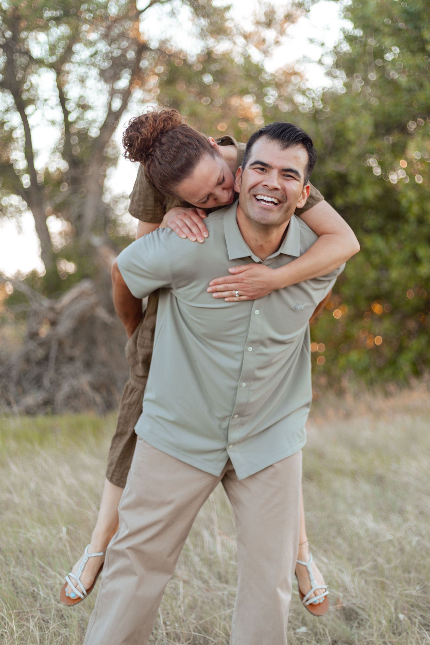 A laughing man gives a woman a piggyback ride in a grassy field, both wearing earth-toned outfits.