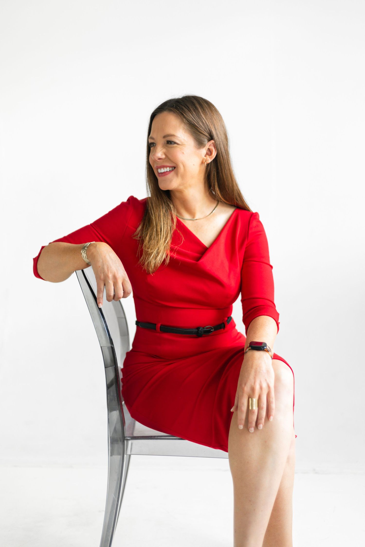Smiling woman in red dress seated on clear chair against white background.