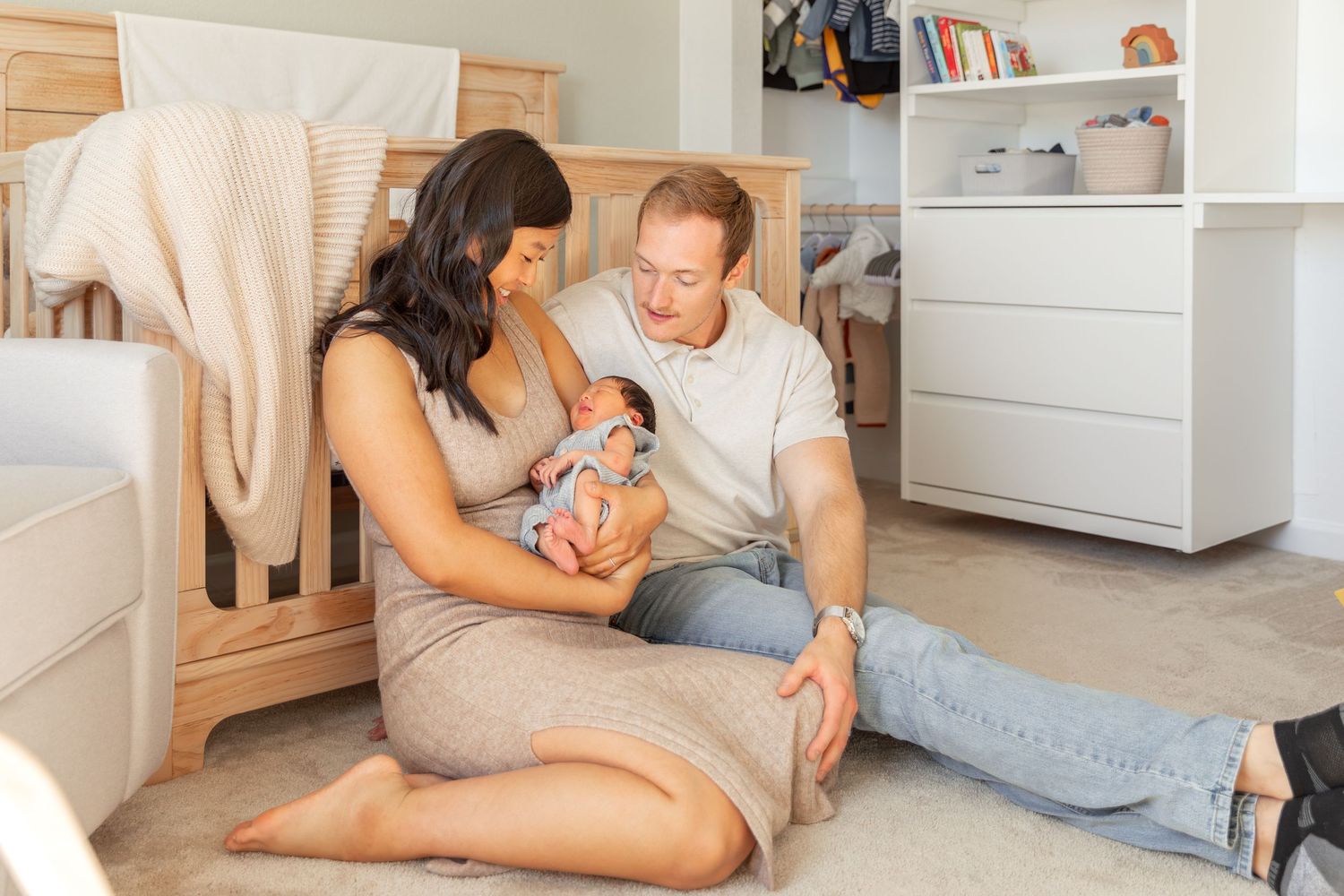 New parents sitting on nursery floor lovingly cradling their newborn baby together near a white crib.