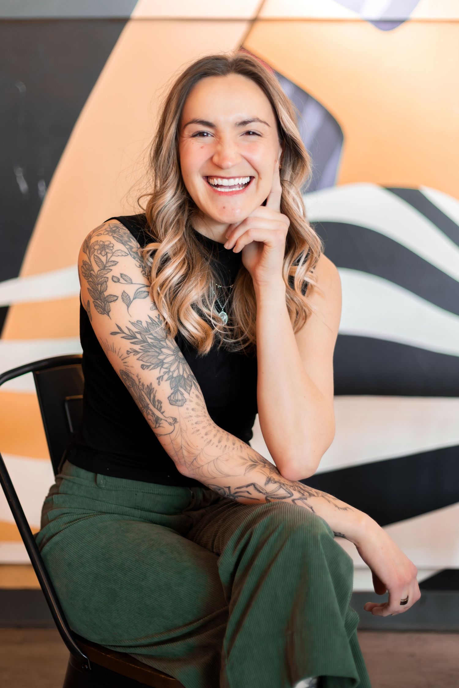 A person in a black top and green pants sits casually in a chair, showing off detailed sleeve tattoo artwork.
