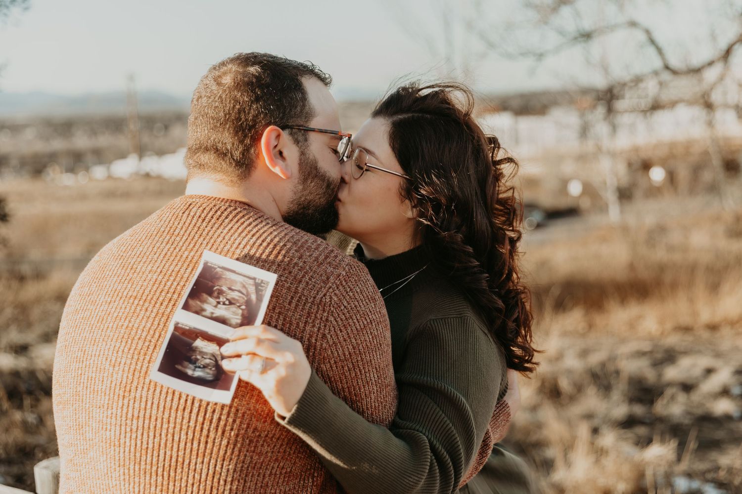 Couple embracing outdoors in warm autumn light while holding ultrasound photos in a tender moment.