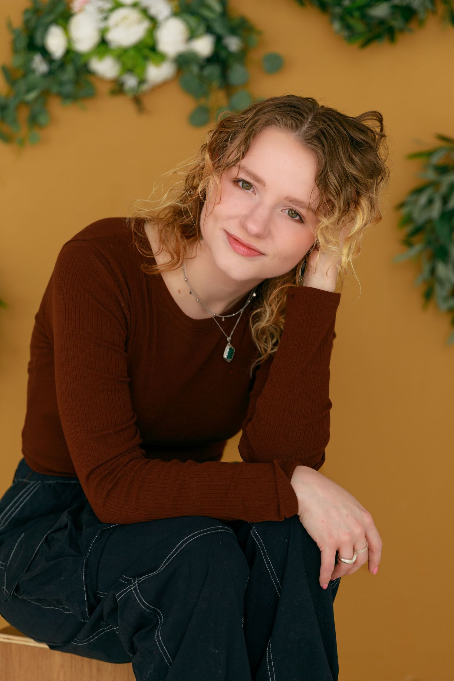 Woman in brown sweater posing against golden wall with greenery decorations.