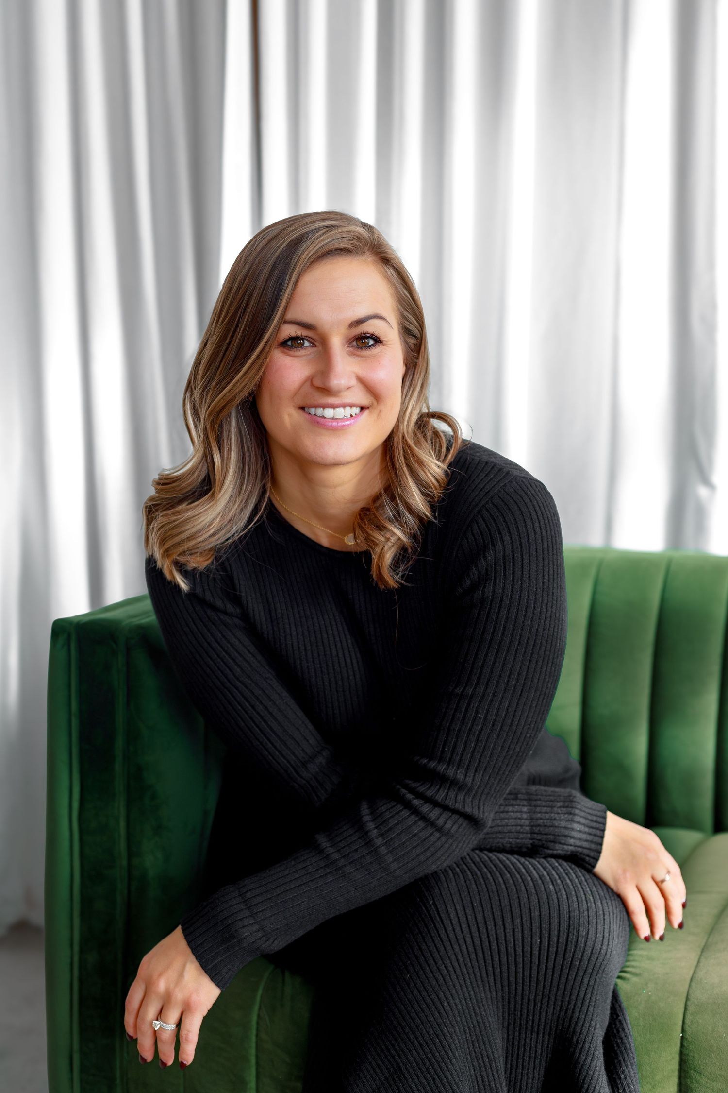 Portrait of someone in black sweater sitting on emerald green velvet chair against white curtain backdrop.