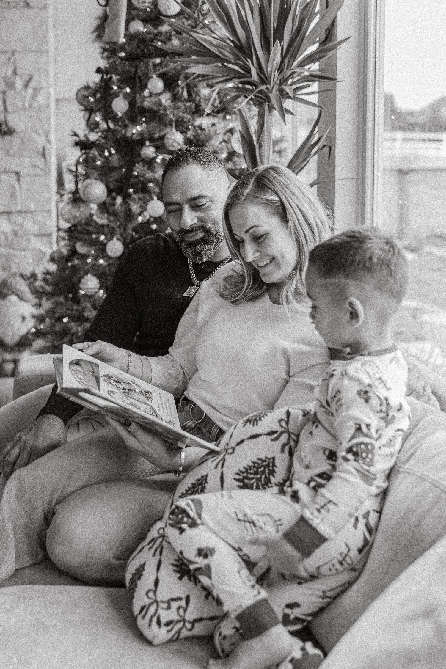 Black and white photo of a family reading a book together on a couch near a decorated Christmas tree.