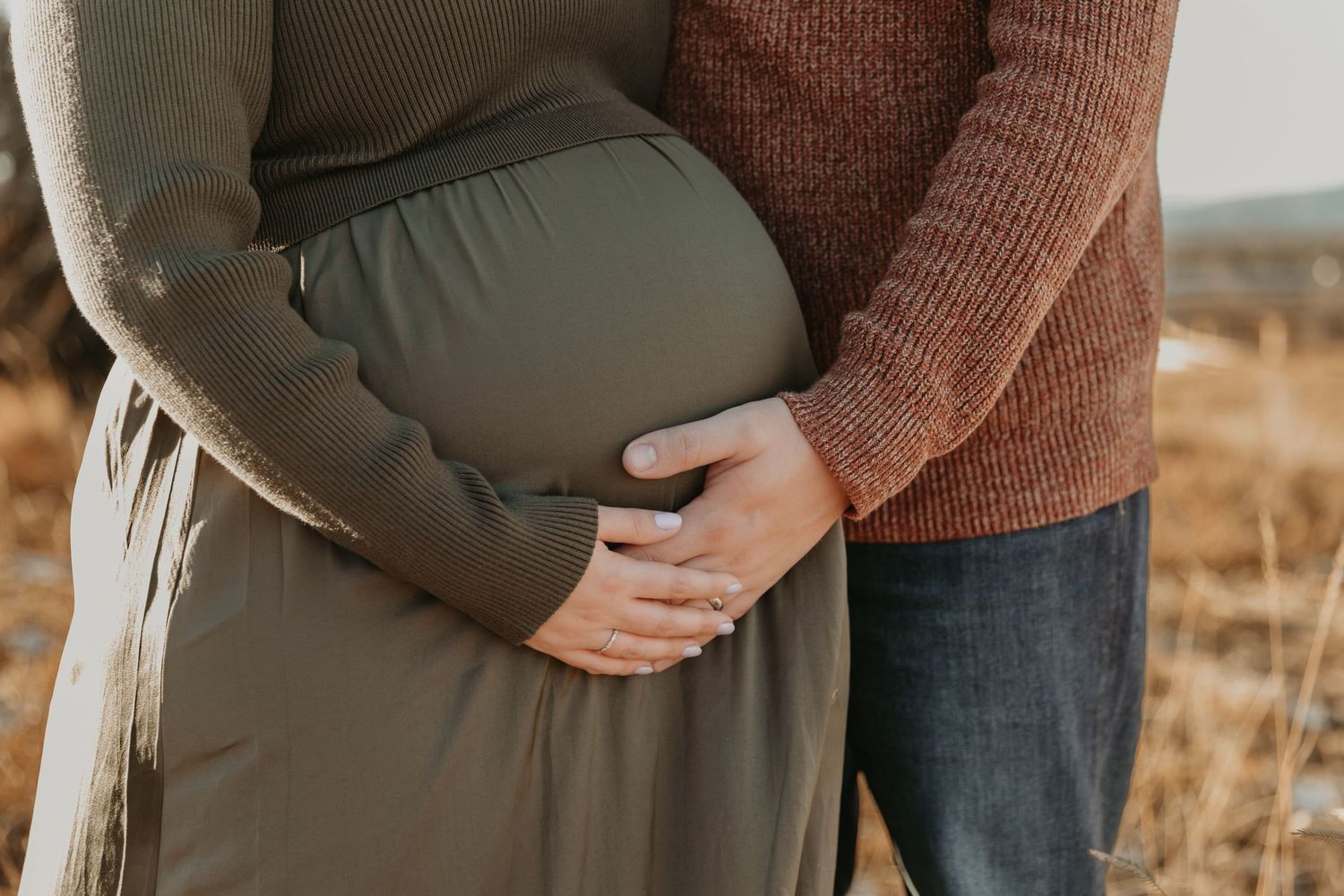 A close-up shot of hands gently cradling a pregnant belly in an olive green dress against a warm autumn background.