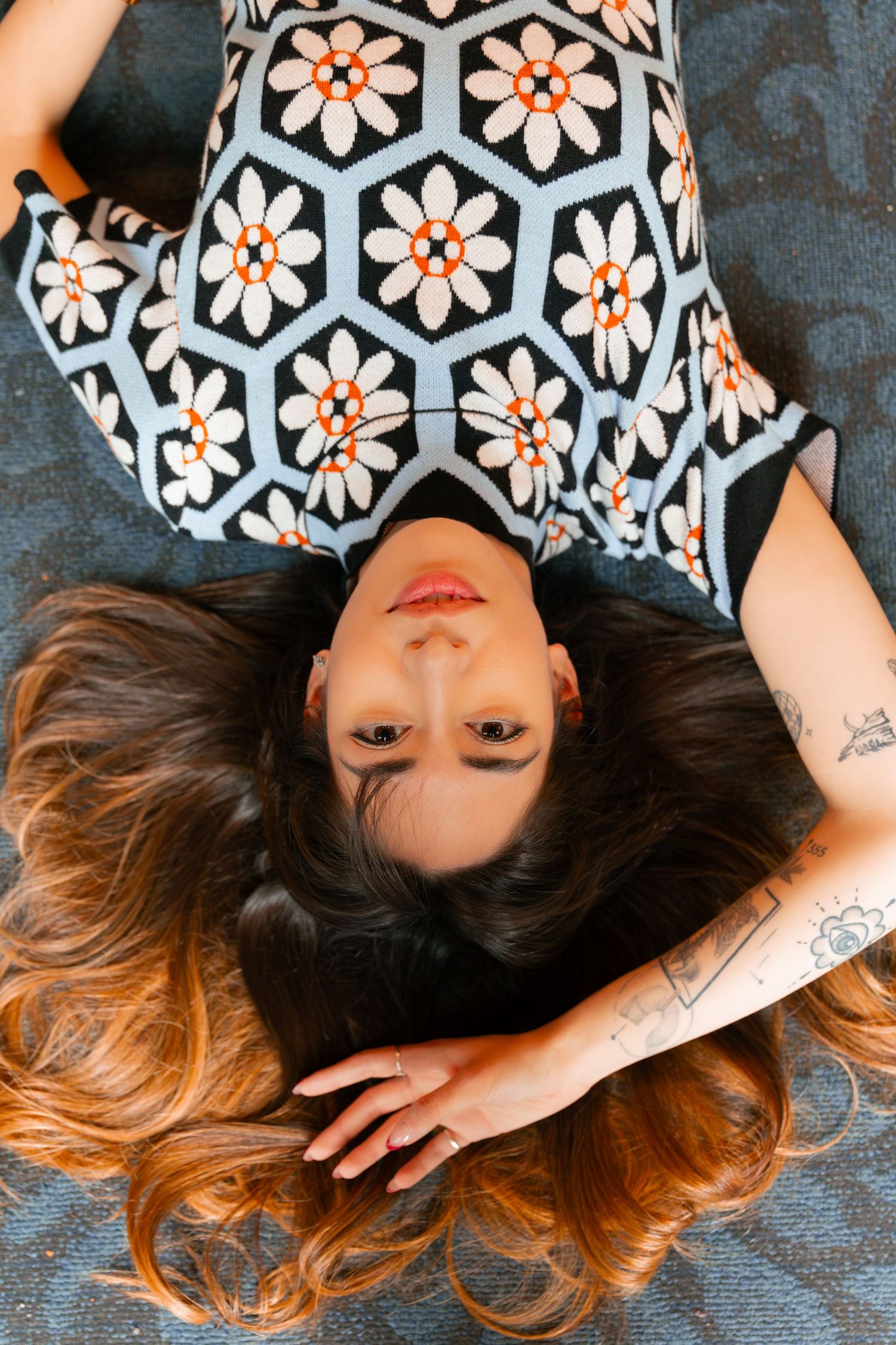 Overhead view of person in floral dress lying on blue carpet near bookshelves.