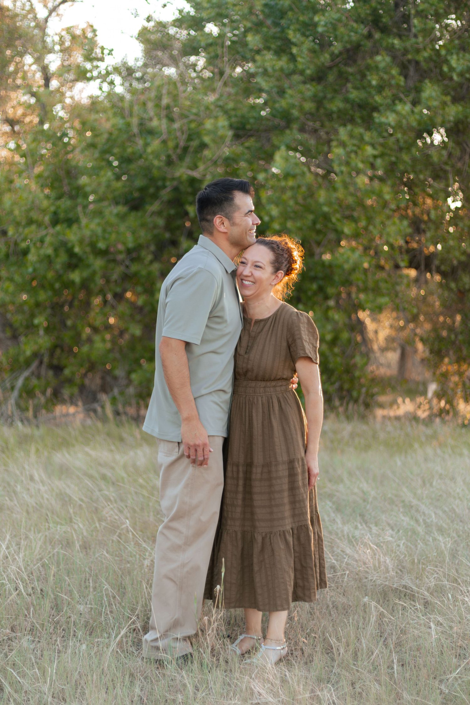 Couple embracing in a golden-lit meadow, woman in brown dress smiling, man in gray shirt, surrounded by green trees.