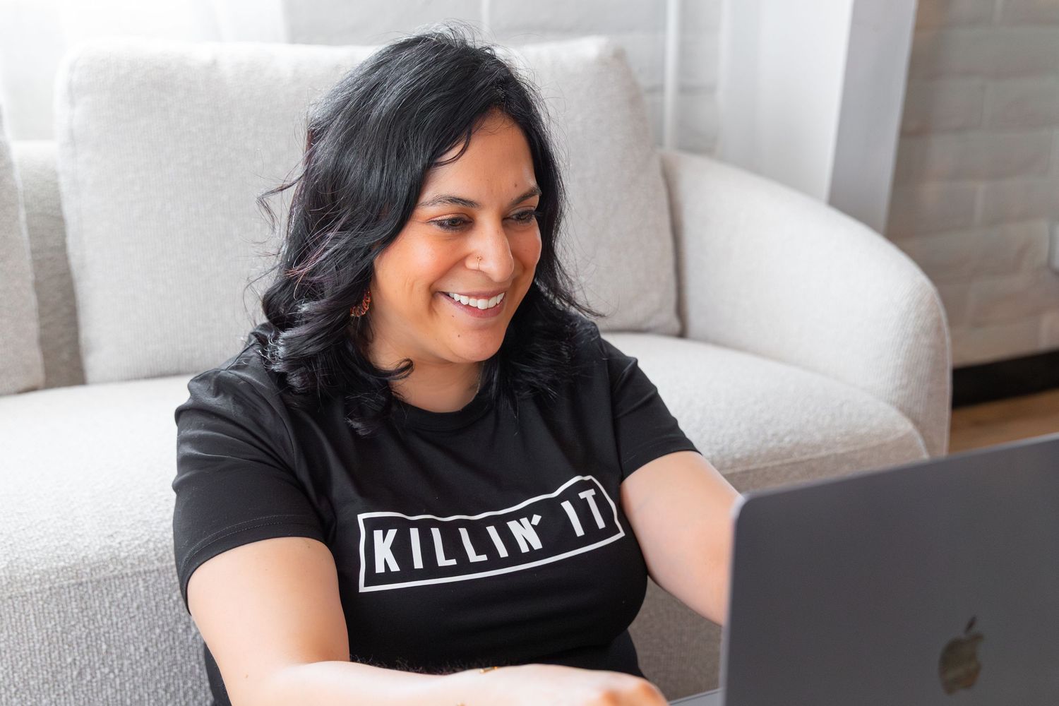 A person wearing a 'Killin' It' t-shirt smiles while working on their MacBook.