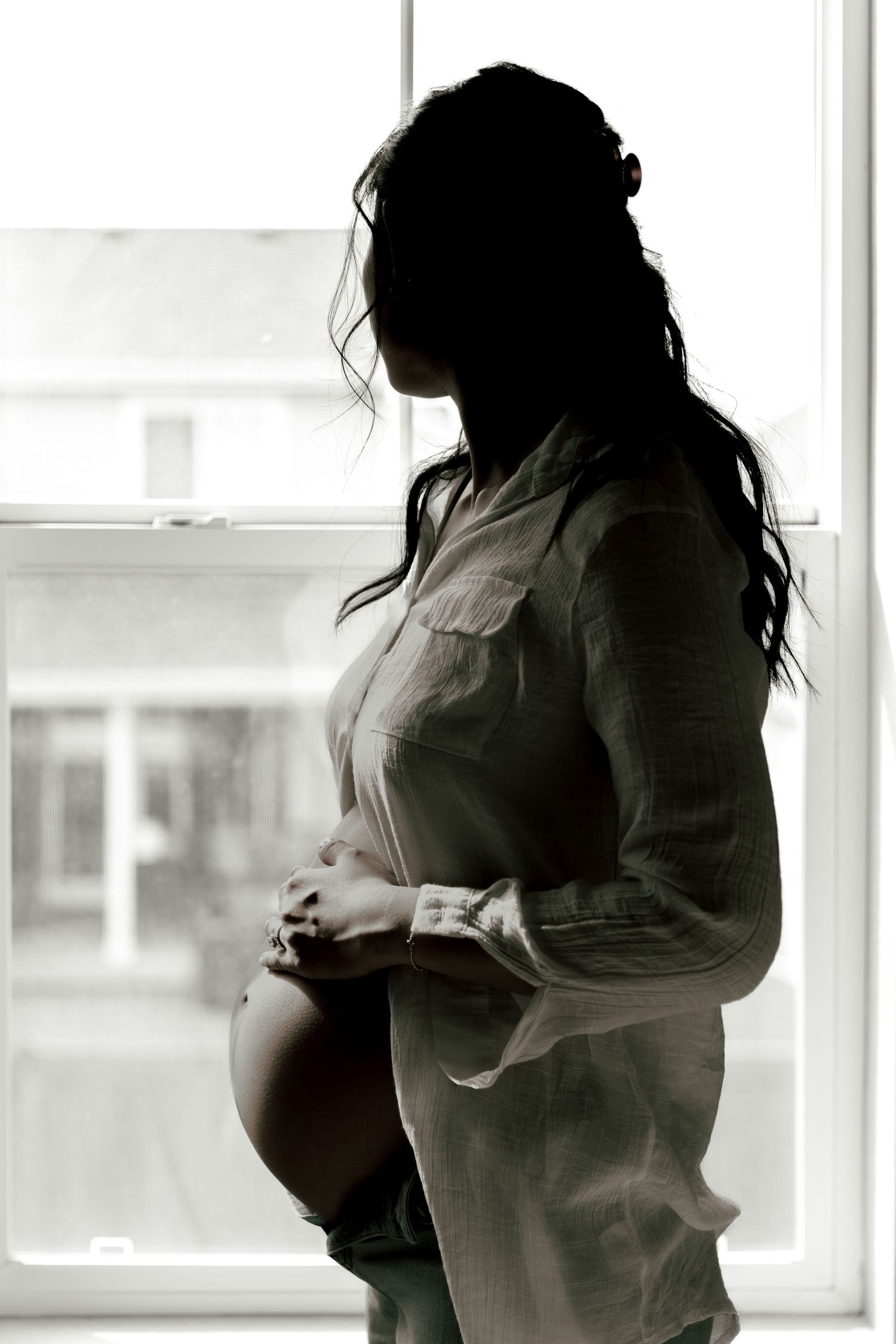 Black and white silhouette of pregnant woman in button-down shirt standing by window in contemplative pose.