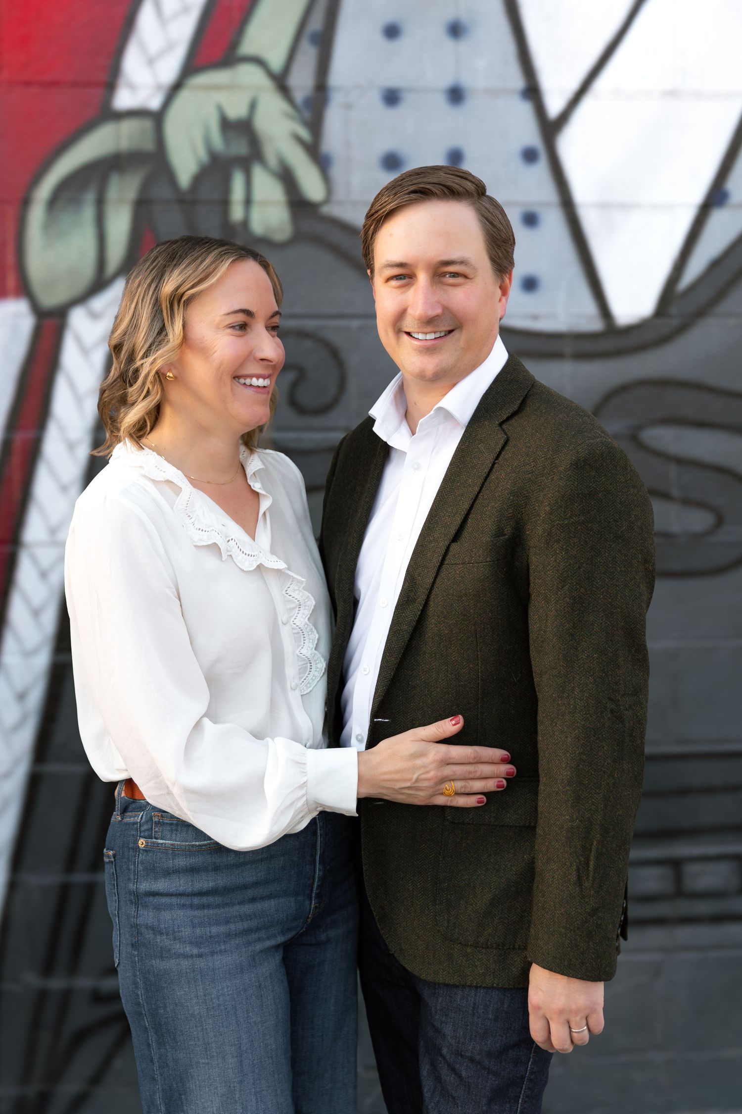 A couple poses against a graffiti wall, wearing casual business attire with a white blouse and dark blazer.