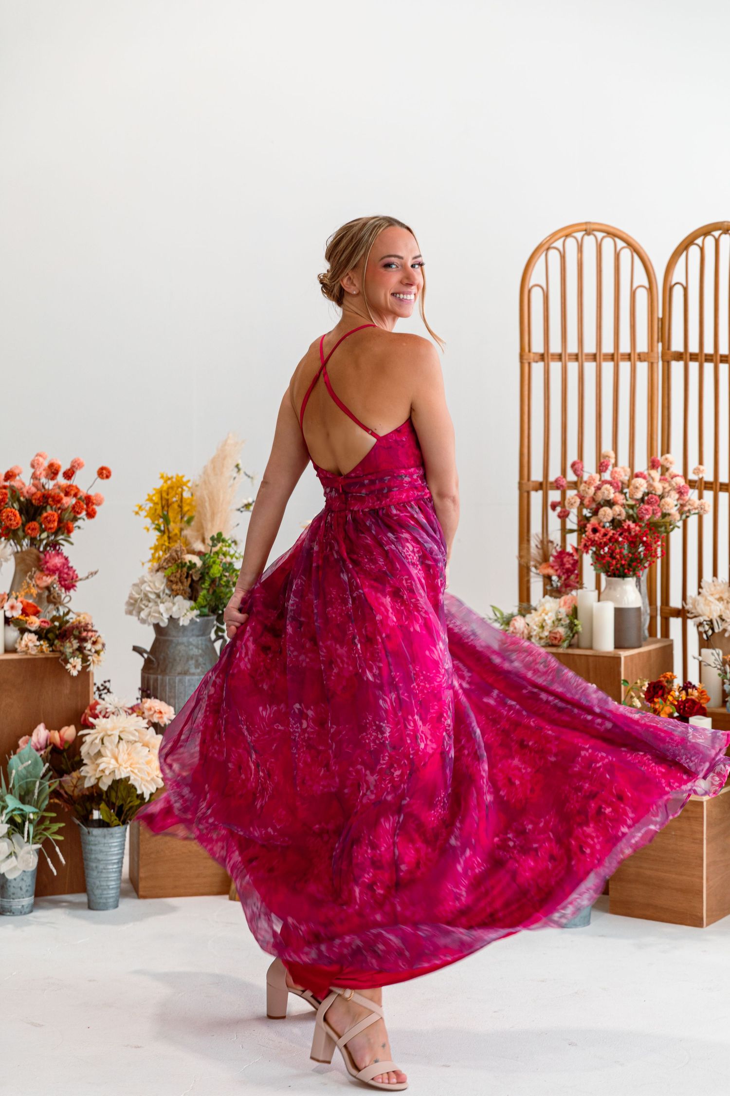 Flowing magenta lace dress with criss-cross back straps twirls elegantly among floral arrangements and rattan divider.