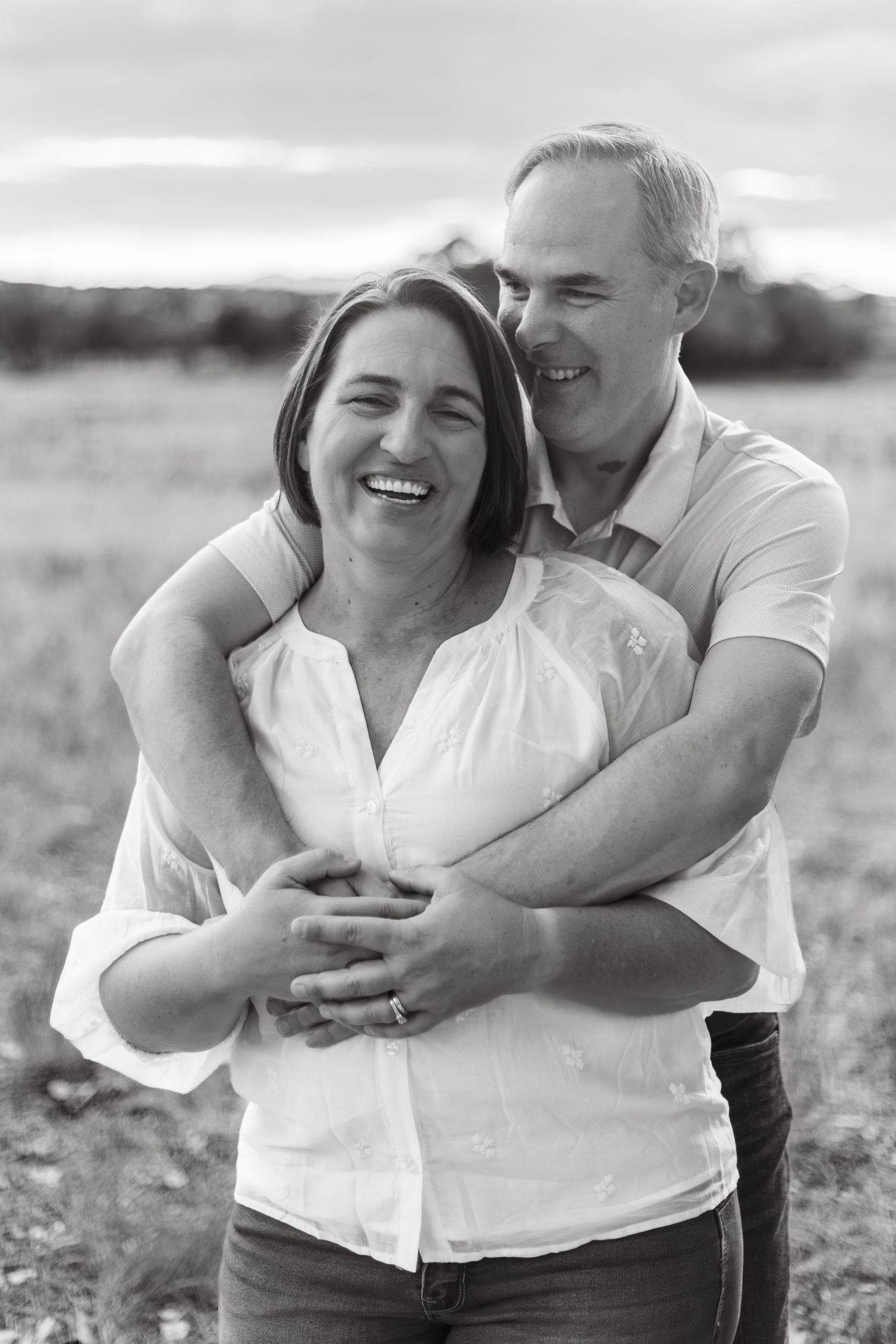 Black and white photo of a happy couple embracing outdoors, man hugging woman from behind as she laughs joyfully.