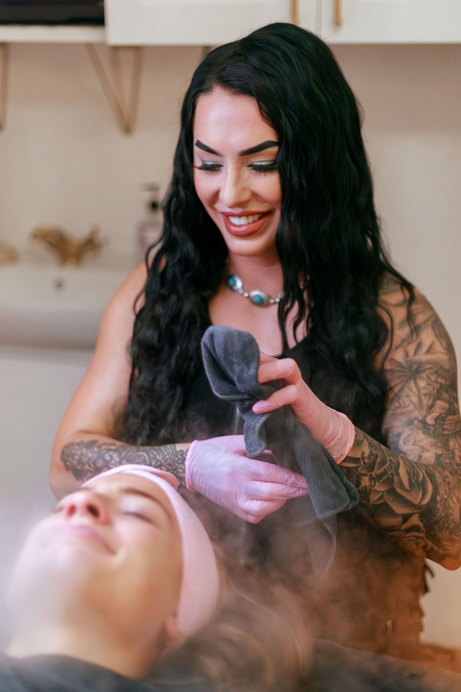 Tattooed esthetician performing facial treatment with steam in spa setting.